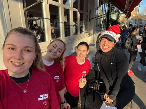 Four young women standing outdoors in front of a building on a city street, smiling at the camera. One woman is wearing a Christmas hat, and another is holding a water bottle. They are dressed casually, with three wearing maroon shirts and one in bla