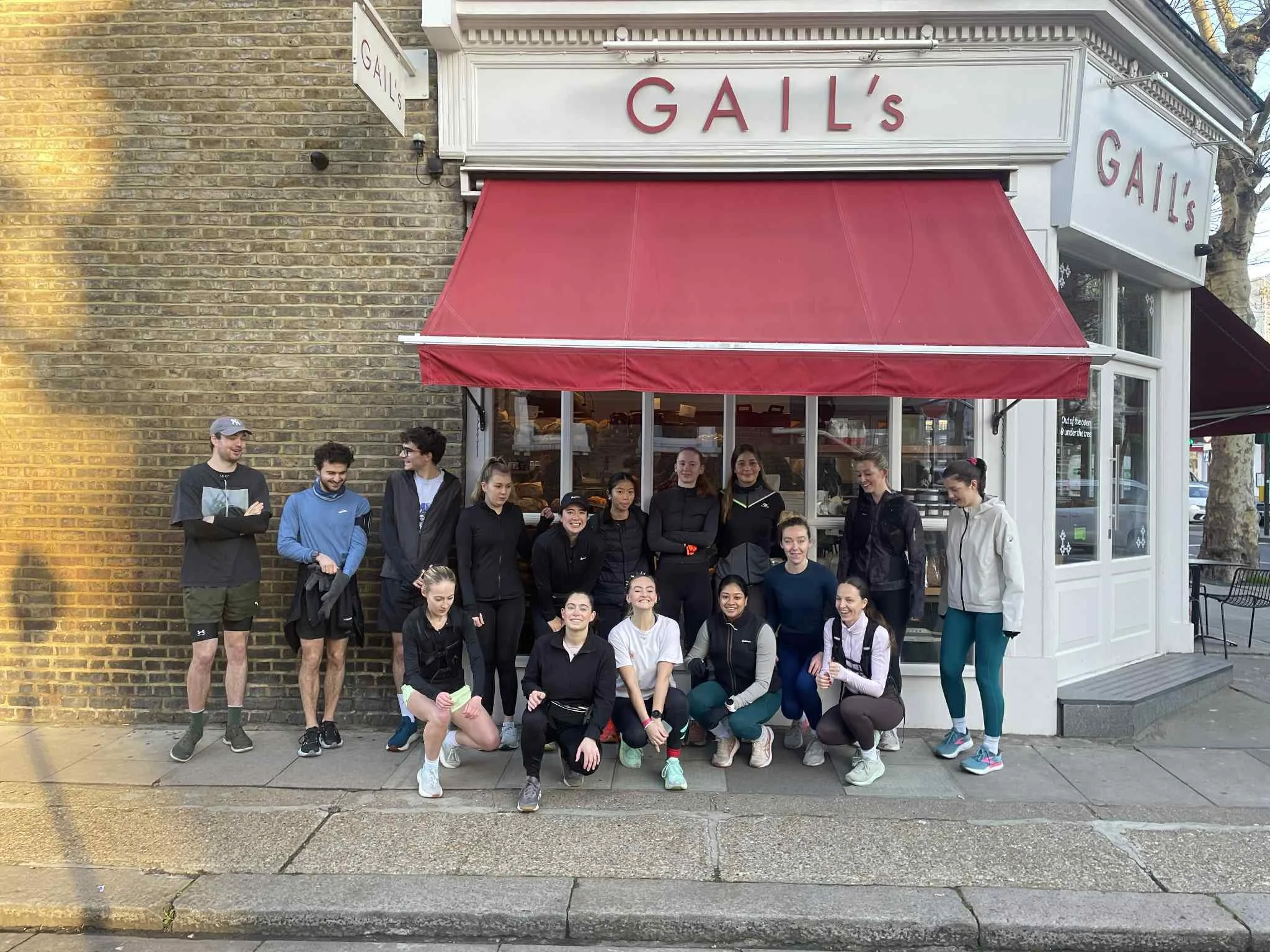 A group of fourteen people, mostly women, standing and kneeling outside a bakery named GAIL'S with a red awning and white storefront. Some are smiling and posing, dressed in athletic or casual clothing.
