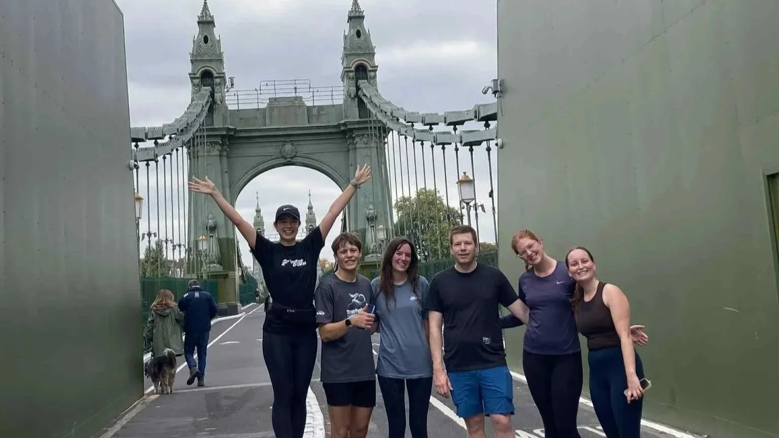Group of six people smiling and posing in front of the Tower Bridge in London, with one person raising arms in celebration.