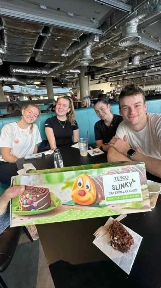 Four young people sitting at a restaurant table, smiling. In the foreground, someone holds a box of Tesco Slinky Caterpillar Cake with a chocolate butterfly flavor, and a piece of the cake on a napkin in front.