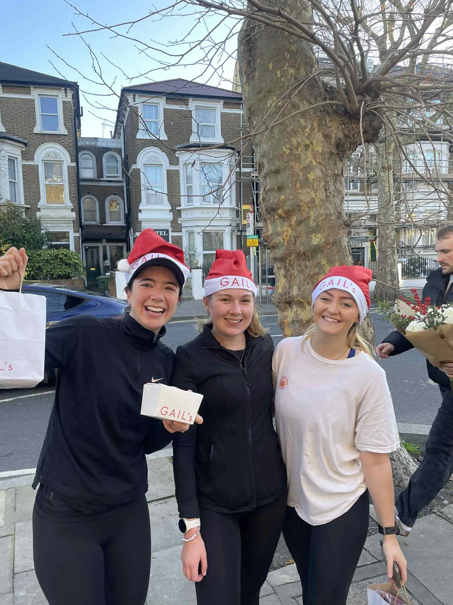 Three women wearing Santa hats with 'GAIL'S' written on them, standing outdoors on a sidewalk with a large tree and row of houses in the background. One woman is holding a gift box and another holding a paper bag. They are smiling.