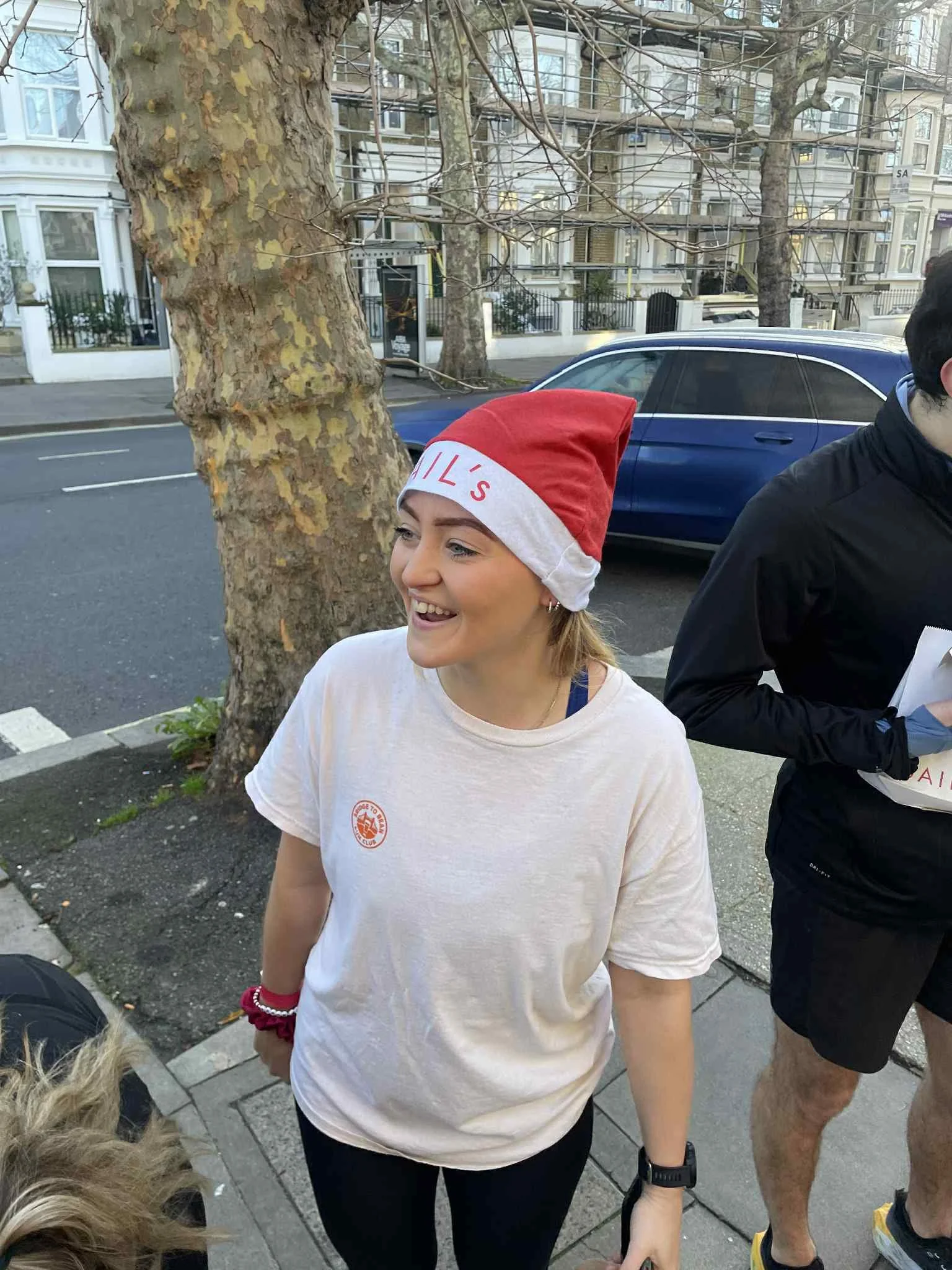 A young woman with blonde hair smiling, wearing a red and white Santa hat that says 'GILL'S', a white T-shirt with a small logo on the chest, black leggings, a black watch, and red and white bracelets, standing on a sidewalk near a tree and a parked 