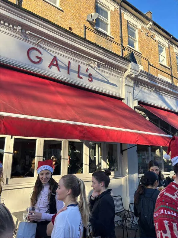 People gathered outside a restaurant named GAIL'S with red awnings, some wearing Santa hats and holiday sweaters.