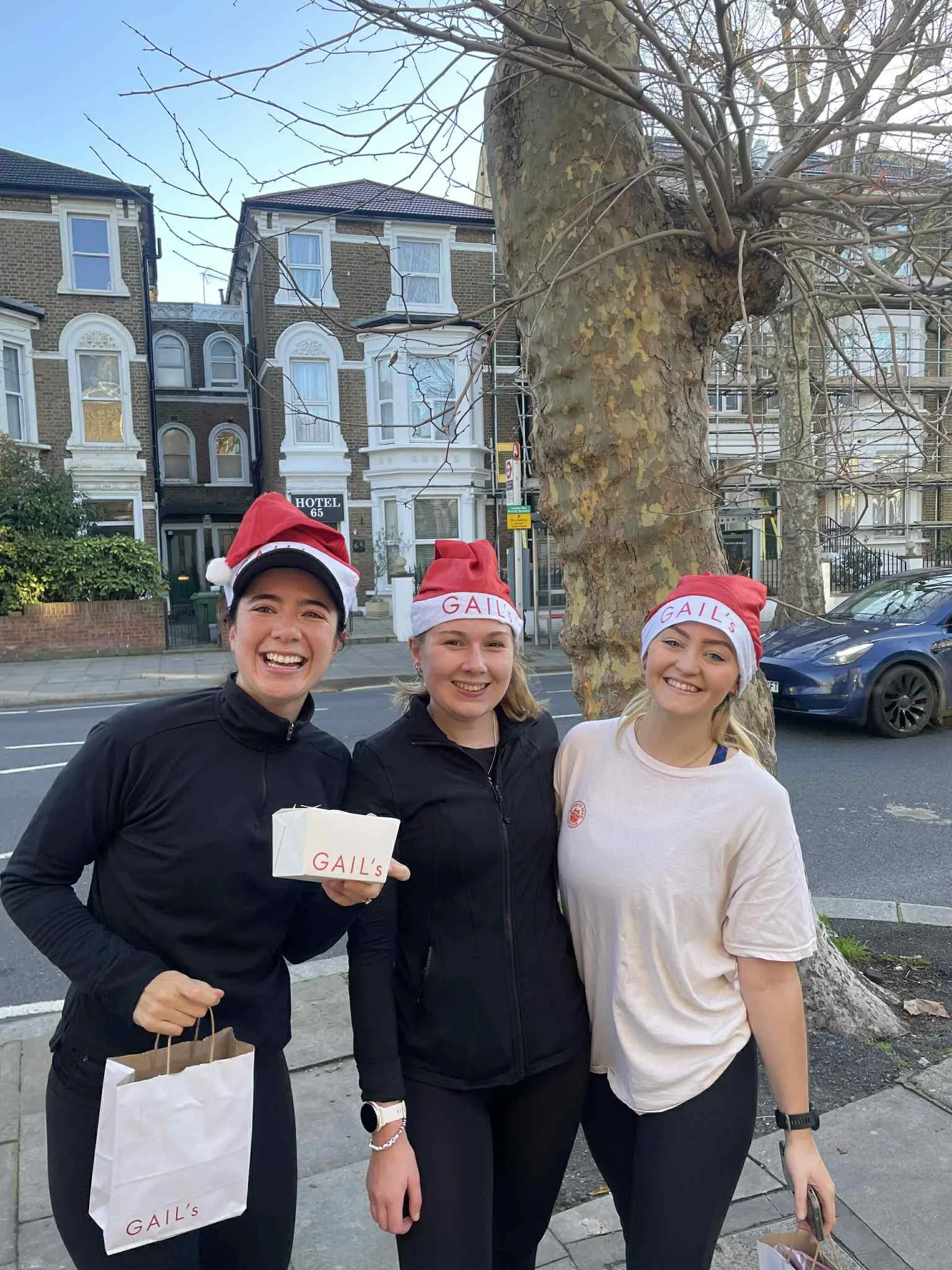 Three women wearing Santa hats with 'GAIL'S' written on them, standing outside on a sidewalk during daytime, smiling.