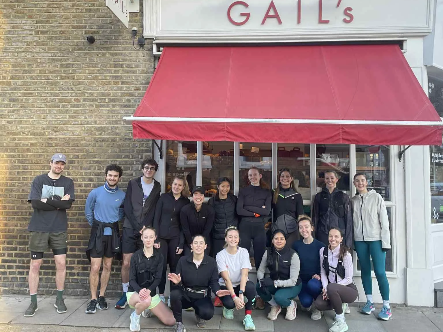 Group of 14 young people in athletic attire standing and kneeling outside a brick building with a red awning, smiling at the camera.