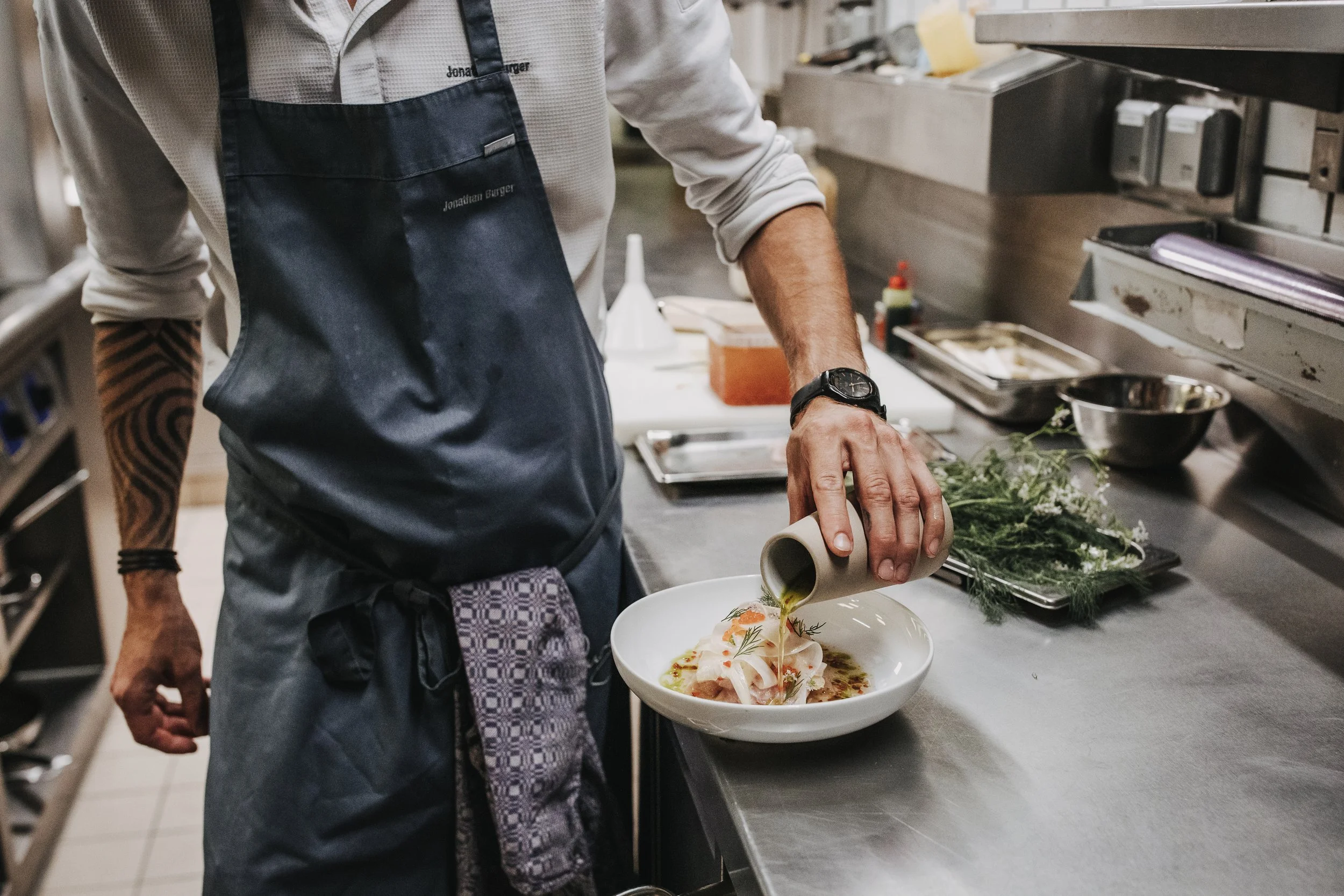 Ein Koch in einer Küche gießt Sauce über ein Gericht in einer Schüssel, umgeben von Küchenutensilien und frischen Kräutern.