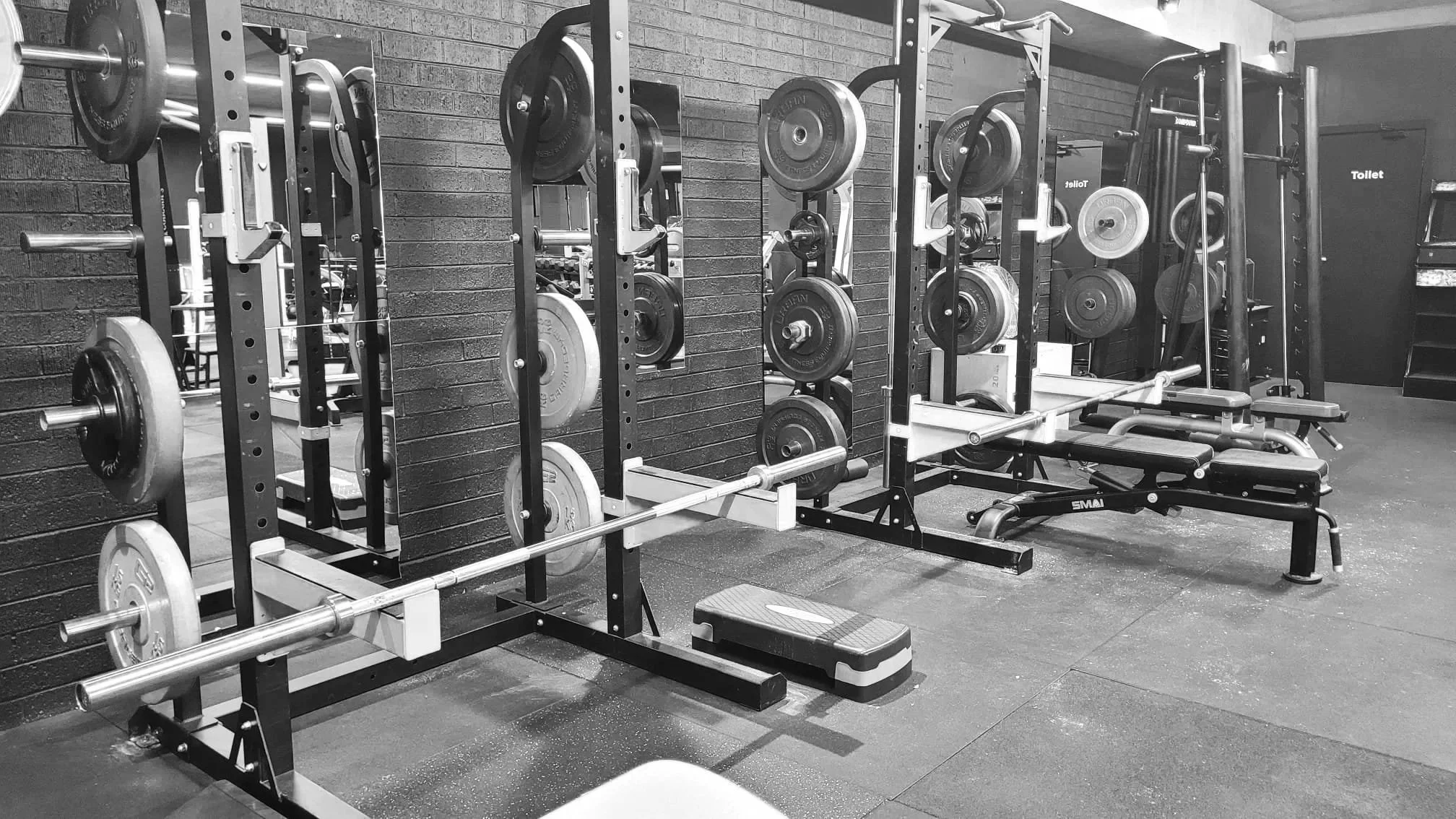 Gym weightlifting area with various weight plates, barbells, and workout equipment against a brick wall.