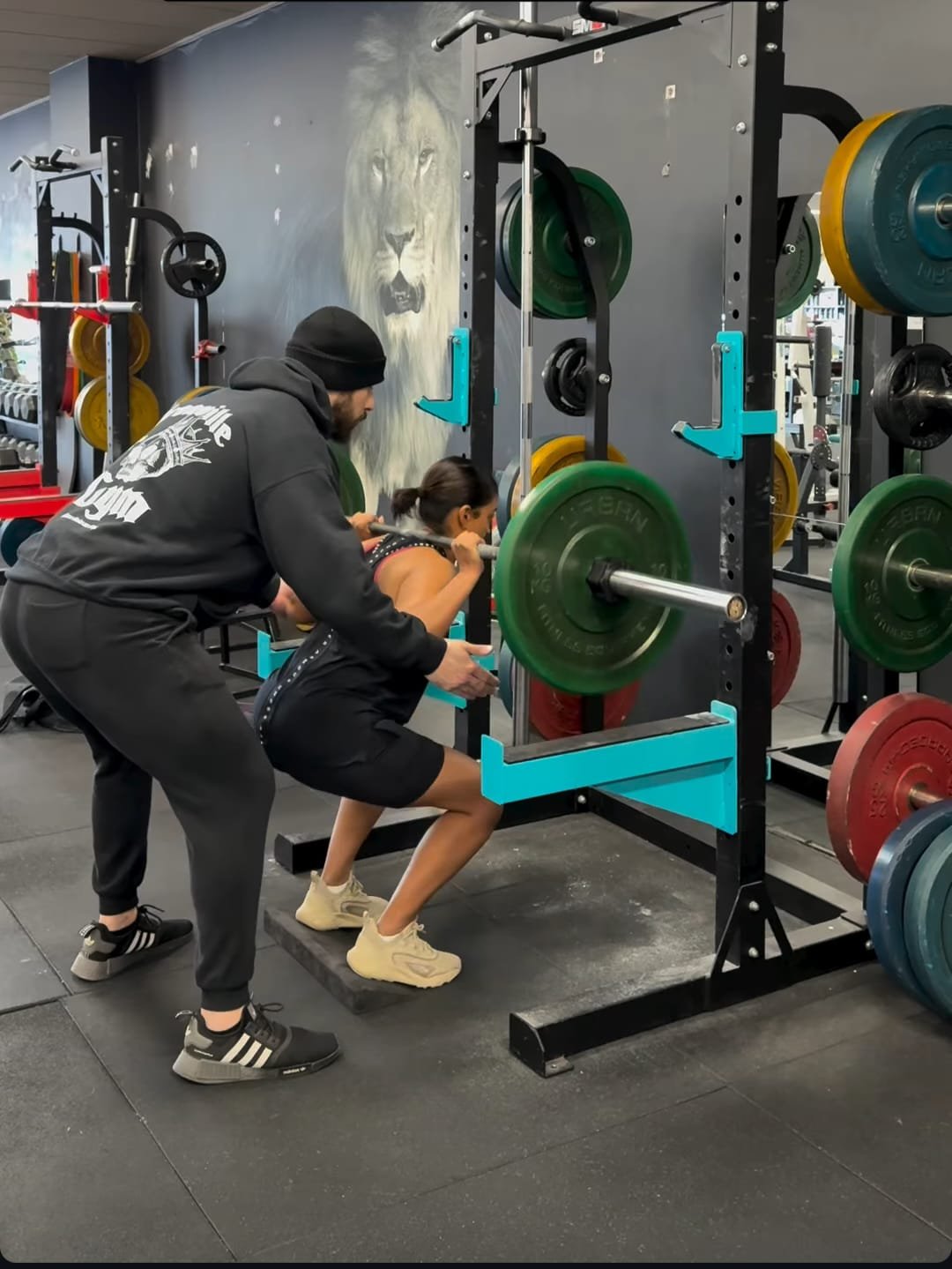 A woman performing a squat exercise with a barbell loaded with weights, supervised by a trainer.