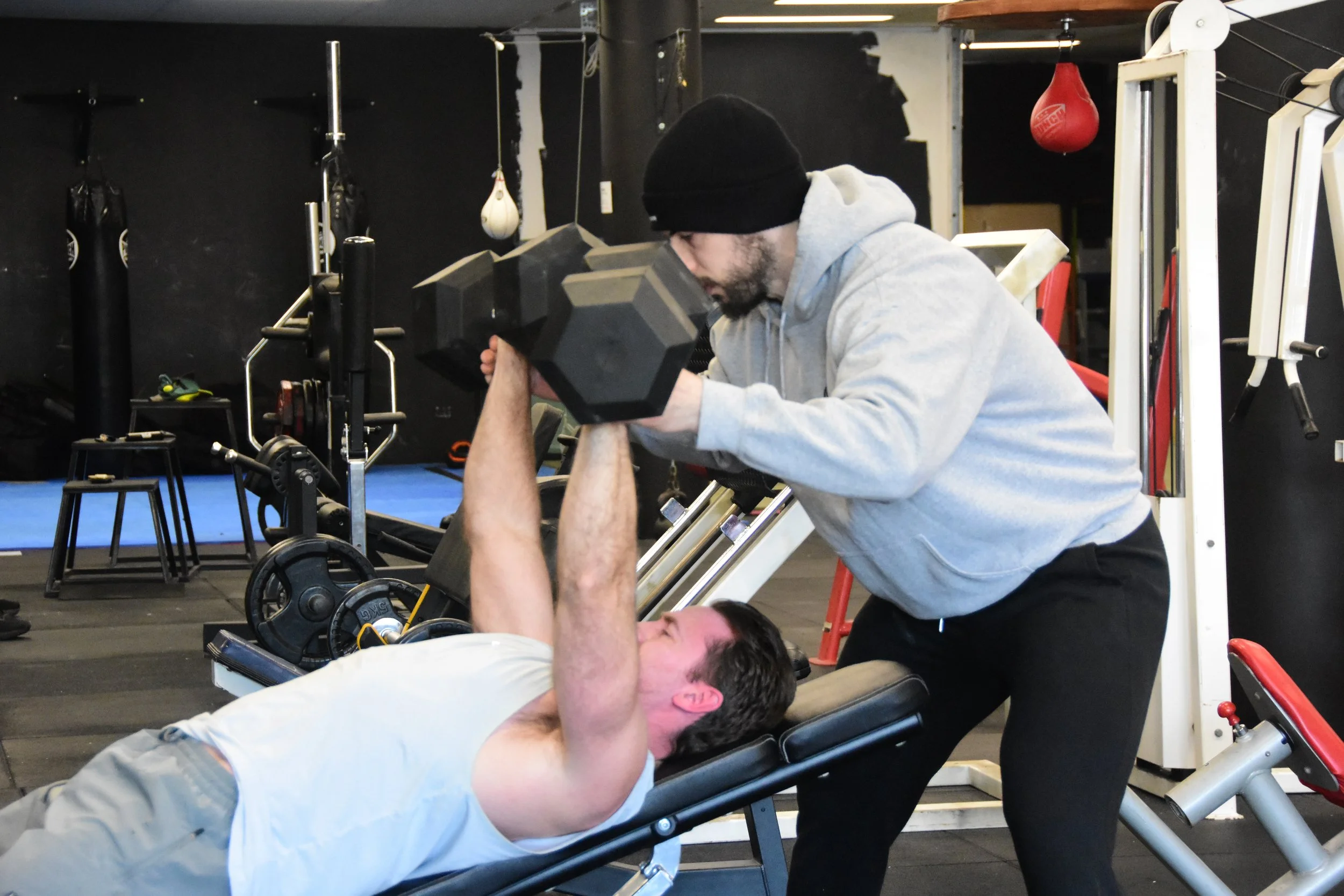 A man lifting a dumbbell while lying on a workout bench at a gym, with a trainer spotting him.