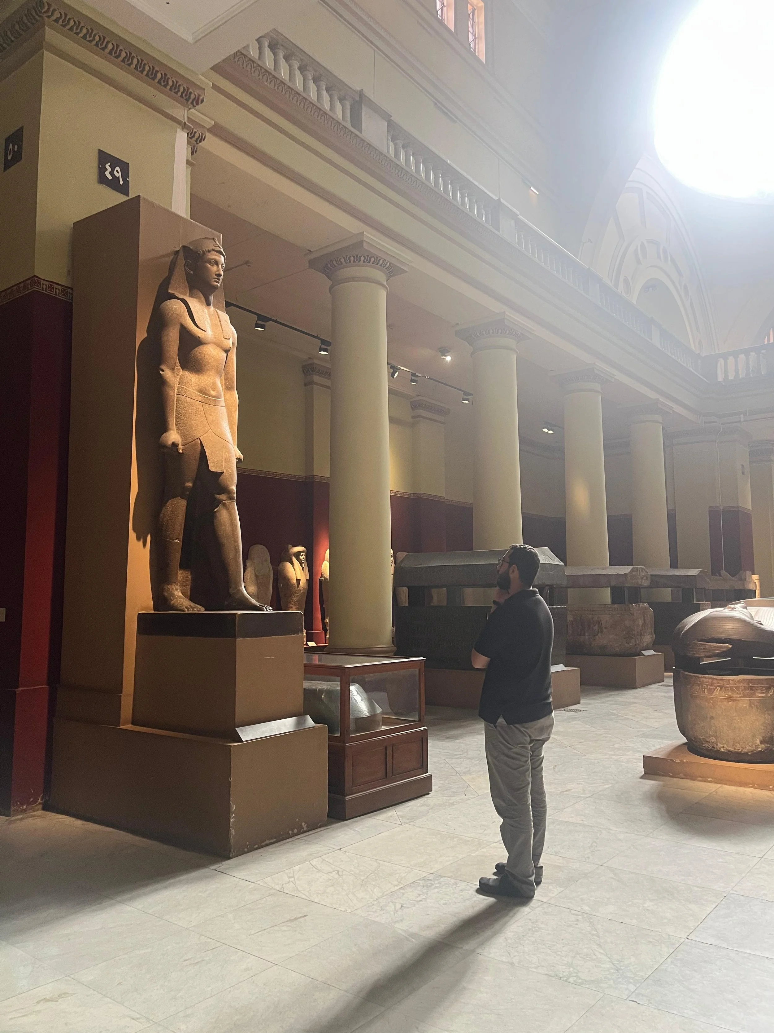 A man stands inside a museum looking at ancient Egyptian sculptures and artifacts. The museum features tall columns and large artifacts displayed around.