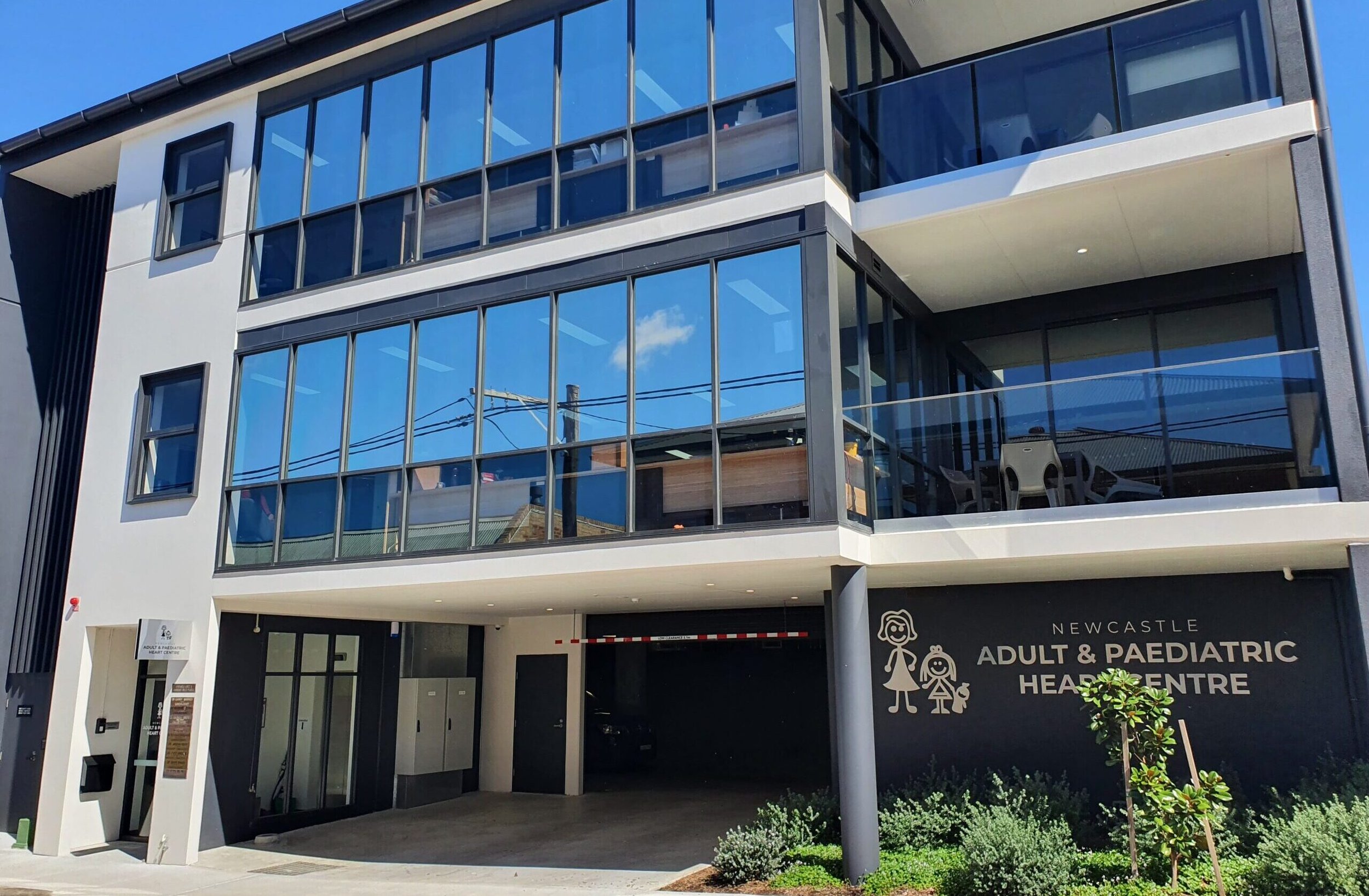Modern multi-story building with large glass windows housing the Newcastle Adult & Paediatric Heart Centre, with signage of a girl, boy, and heart symbol, and greenery in front.