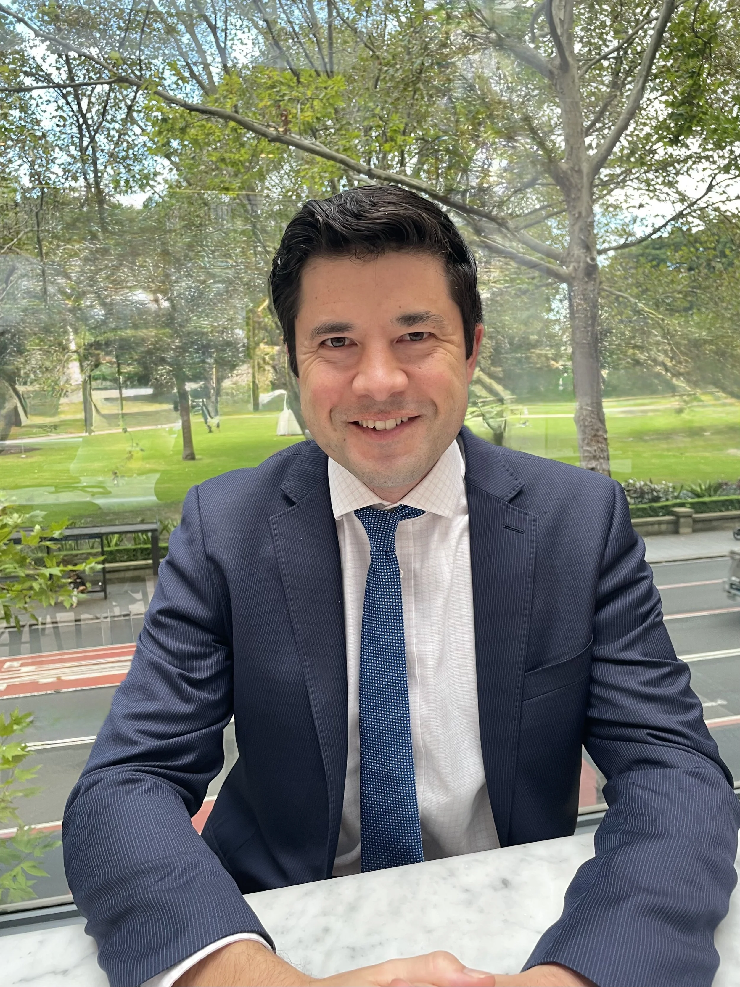 A man in a dark suit and tie smiling at the camera while sitting at a table in a park-like setting with trees and green grass in the background.