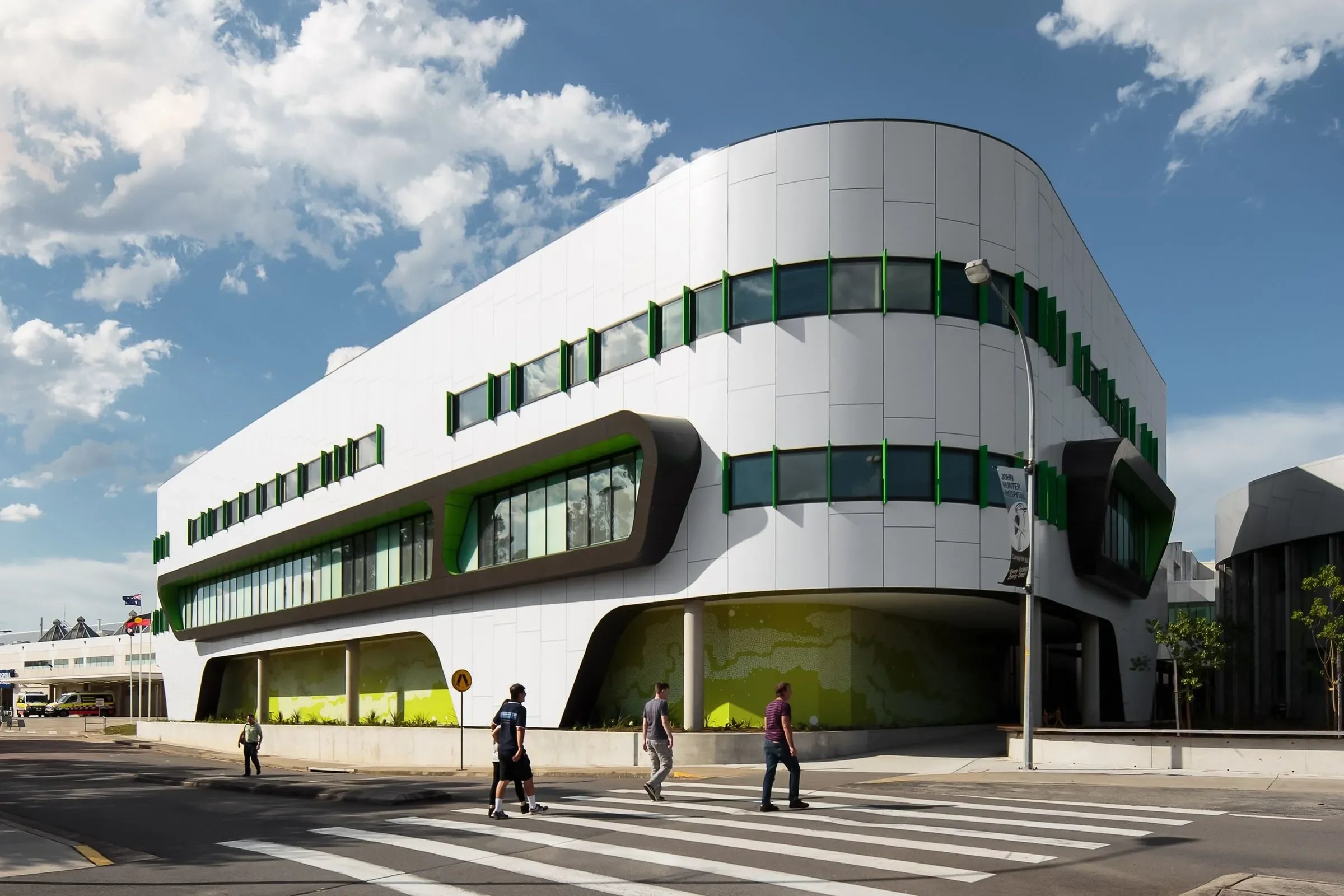 Modern white building with curved architectural design and green accents, situated on a city street with pedestrians crossing the crosswalk under a partly cloudy sky.