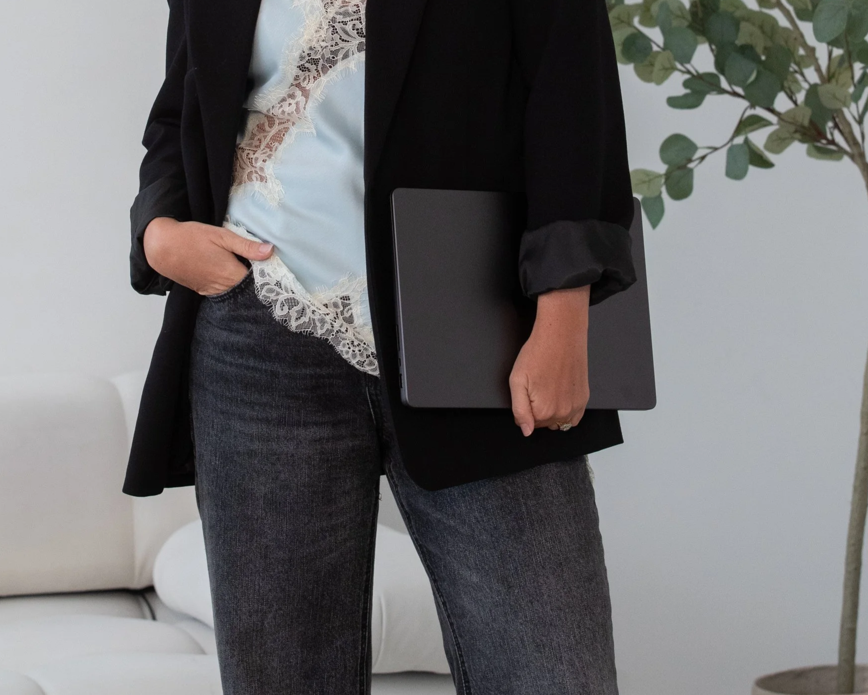 Person wearing a black blazer, teal shirt with lace detailing, and dark jeans, holding a closed black laptop. The background includes a white sofa and a small potted plant.