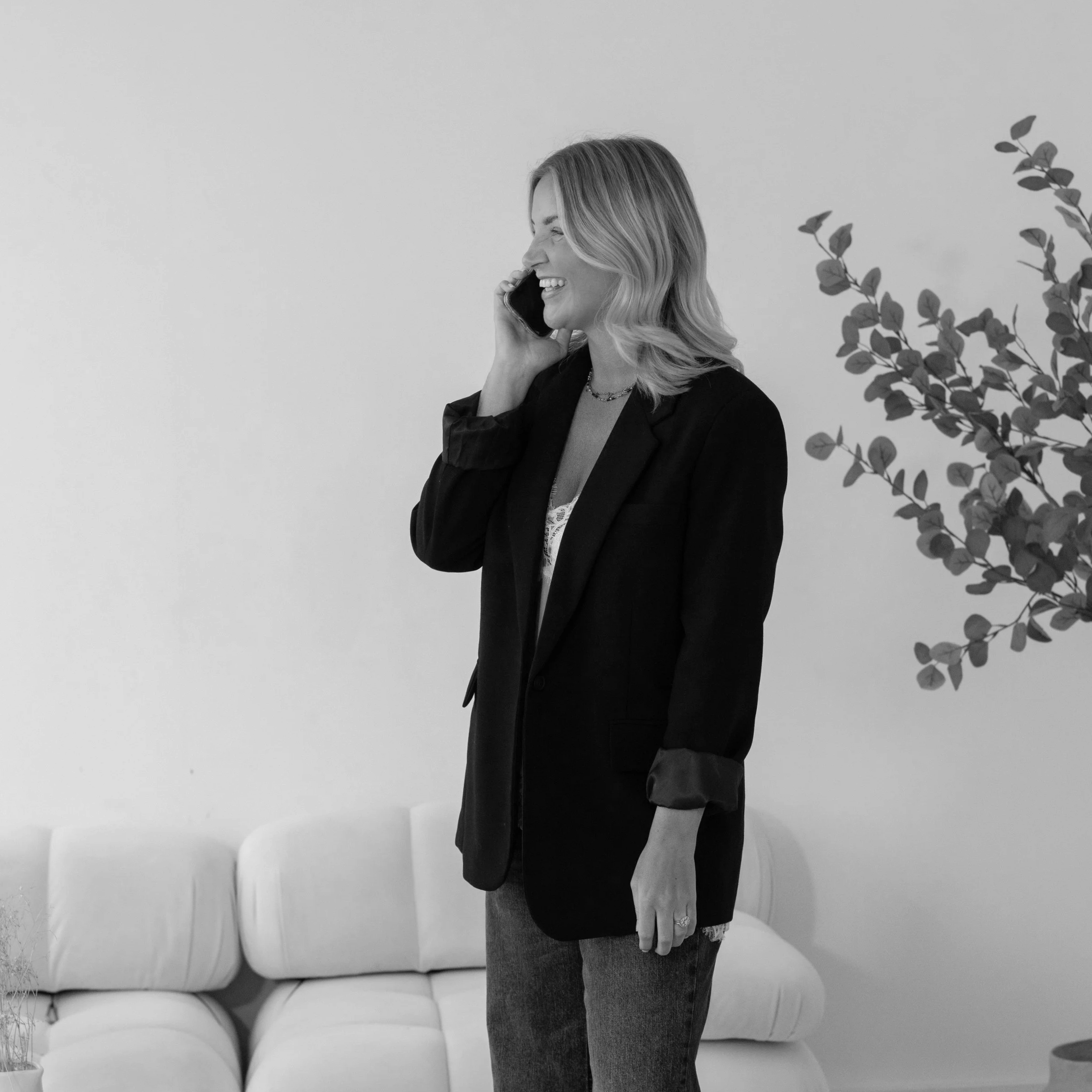 A woman with wavy blonde hair, laughing on the phone in a black blazer and jeans, standing in a modern room with a white couch and potted plant.