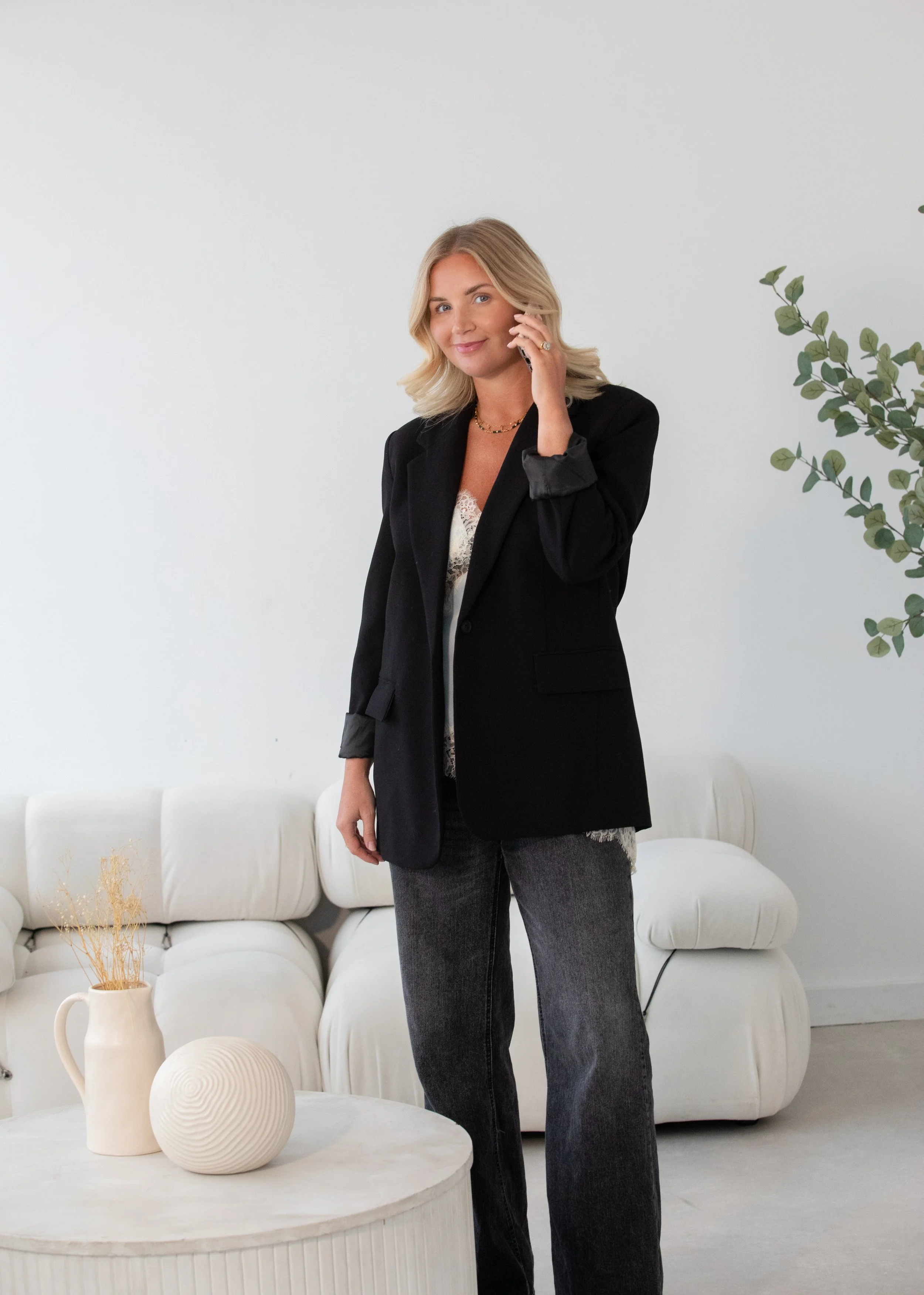 A woman with blonde hair talking on a cell phone in a modern, minimalist living room with white furniture, a beige vase, a spherical decorative object, and a large green plant.