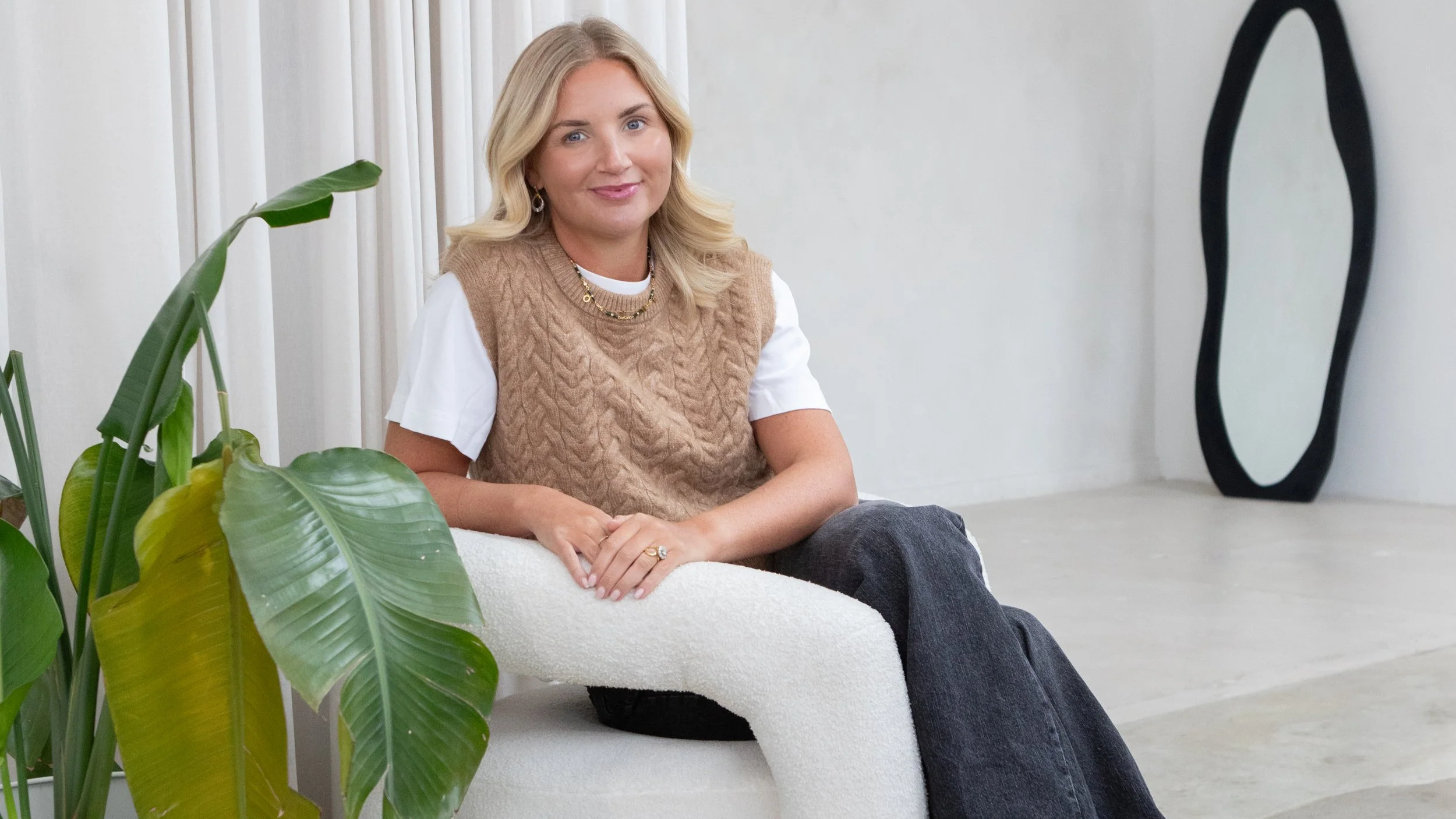 Woman sitting on a modern white armchair in a minimalistic, light-colored room, with a large green plant in the foreground and a black decorative mirror in the background.