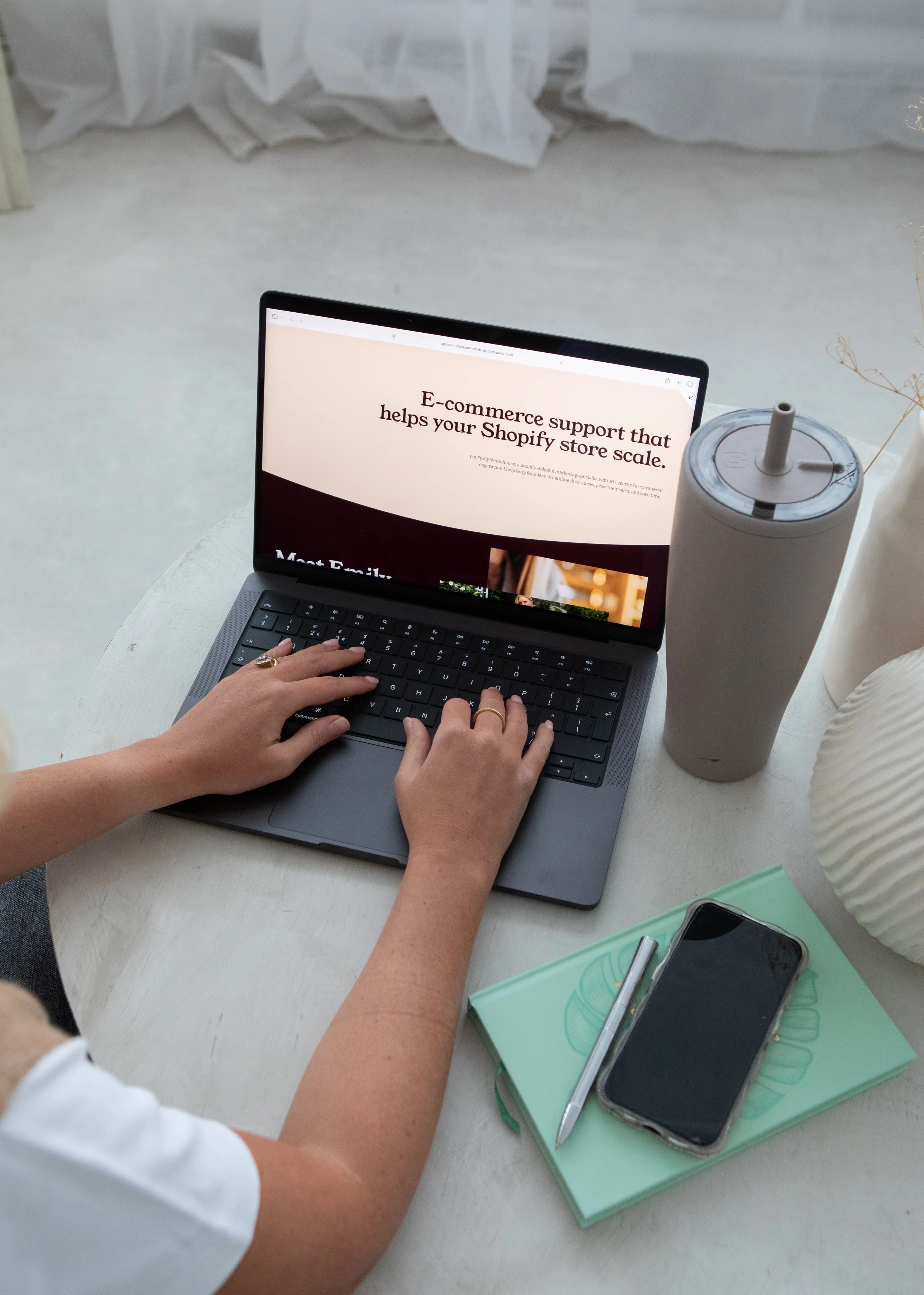 A person working on a laptop computer on a white table, with a green notebook, a pen, and a smartphone nearby. The laptop screen displays a webpage about e-commerce support for Shopify stores. There is also a tall, white tumbler with a straw and a large white vase on the table.
