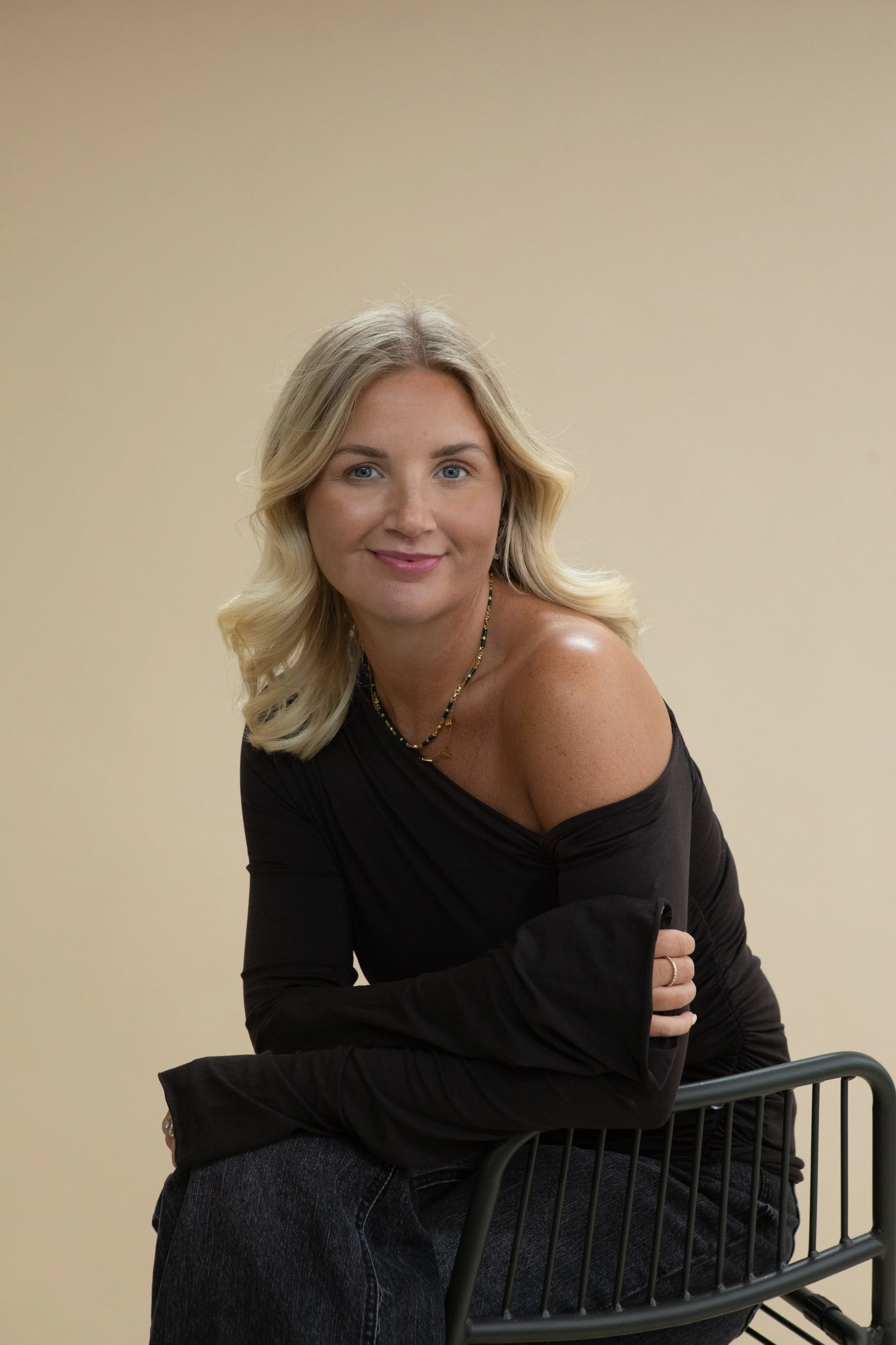 A blonde woman with wavy hair, wearing a black off-shoulder top and jewelry, sitting on a black chair against a plain beige background.
