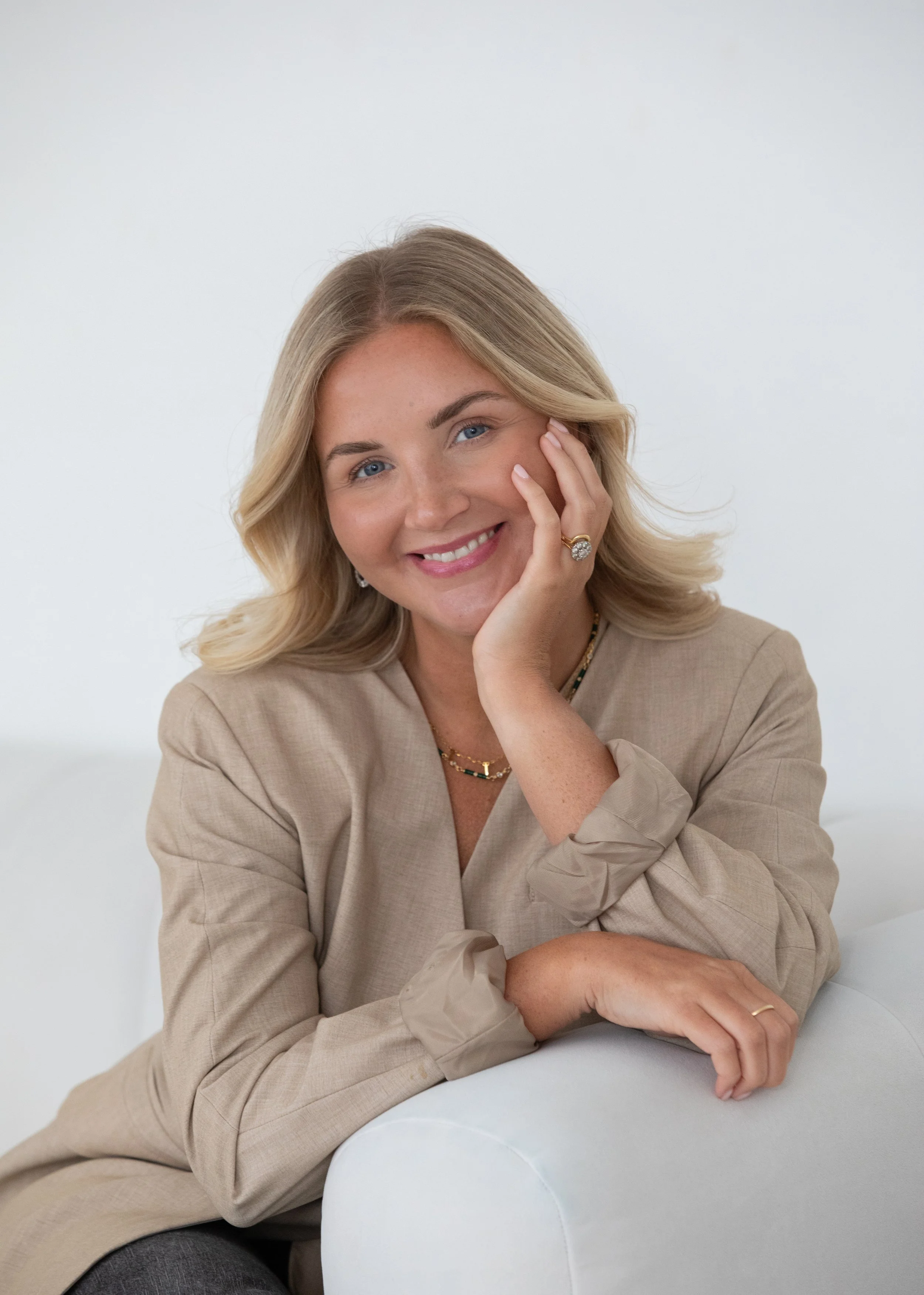 A smiling woman with blonde hair wearing a beige blazer, gold jewelry, and rings, resting her chin on her hand, sitting on a white sofa against a plain white background.