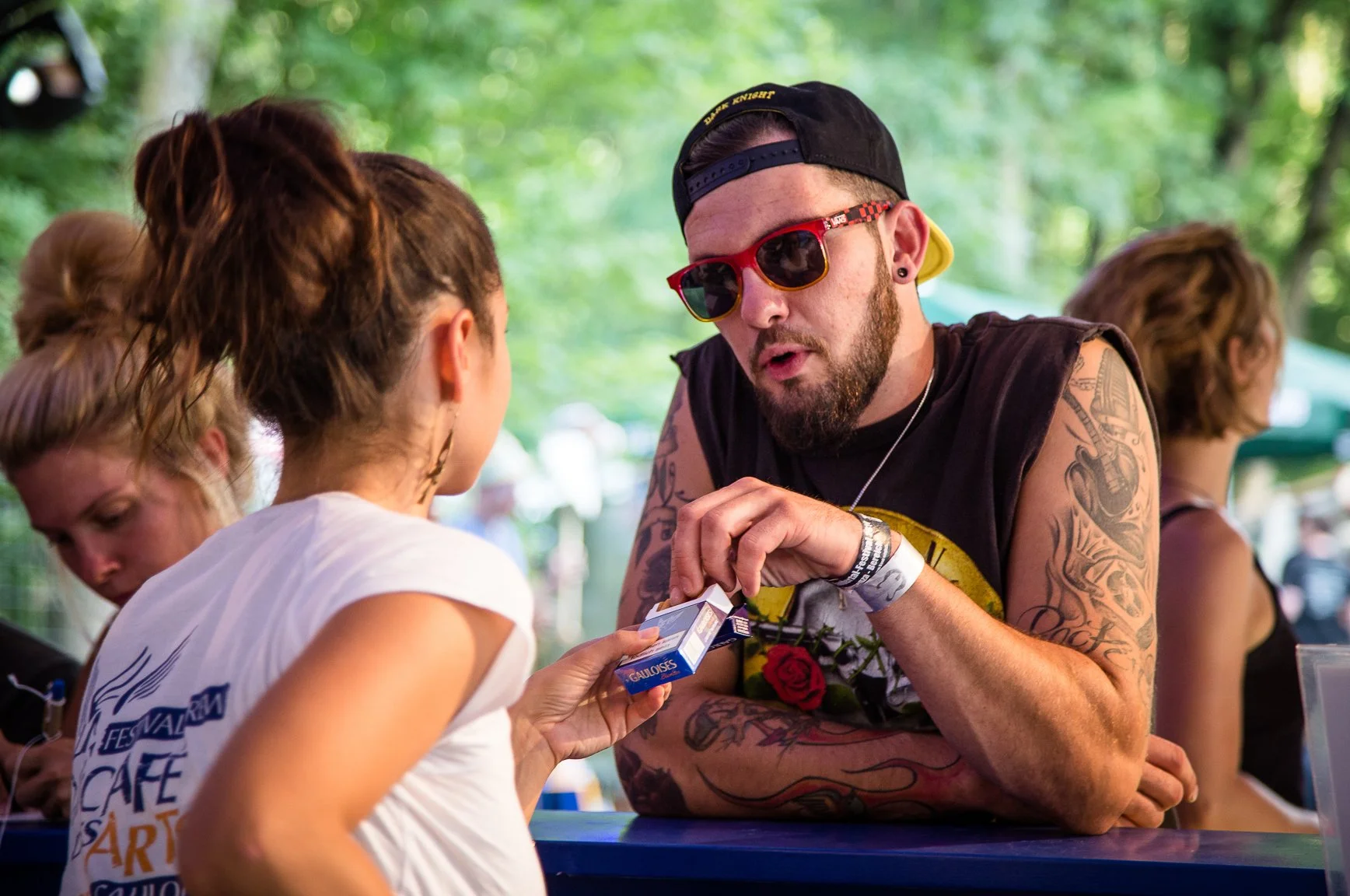 Mann mit Tattoos, roten Sonnenbrillen und schwarzem Cap, der einem jungen Mädchen Zigaretten verkauft auf einem Festival im Freien.