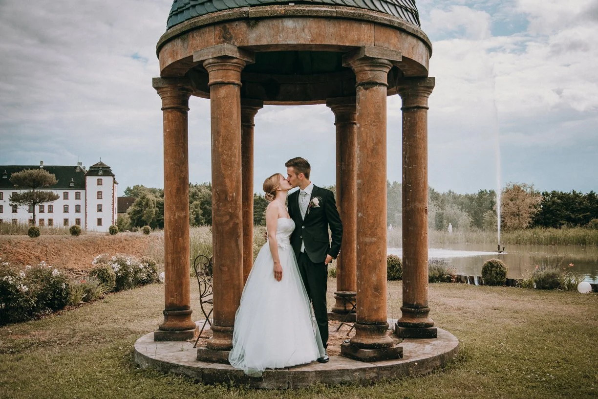 Ein Brautpaar küsst sich unter einem kleinen runden Pavillon mit Säulen, im Freien vor einem Wasserreservoir mit Springbrunnen, im Hintergrund ein großes Schloss.
