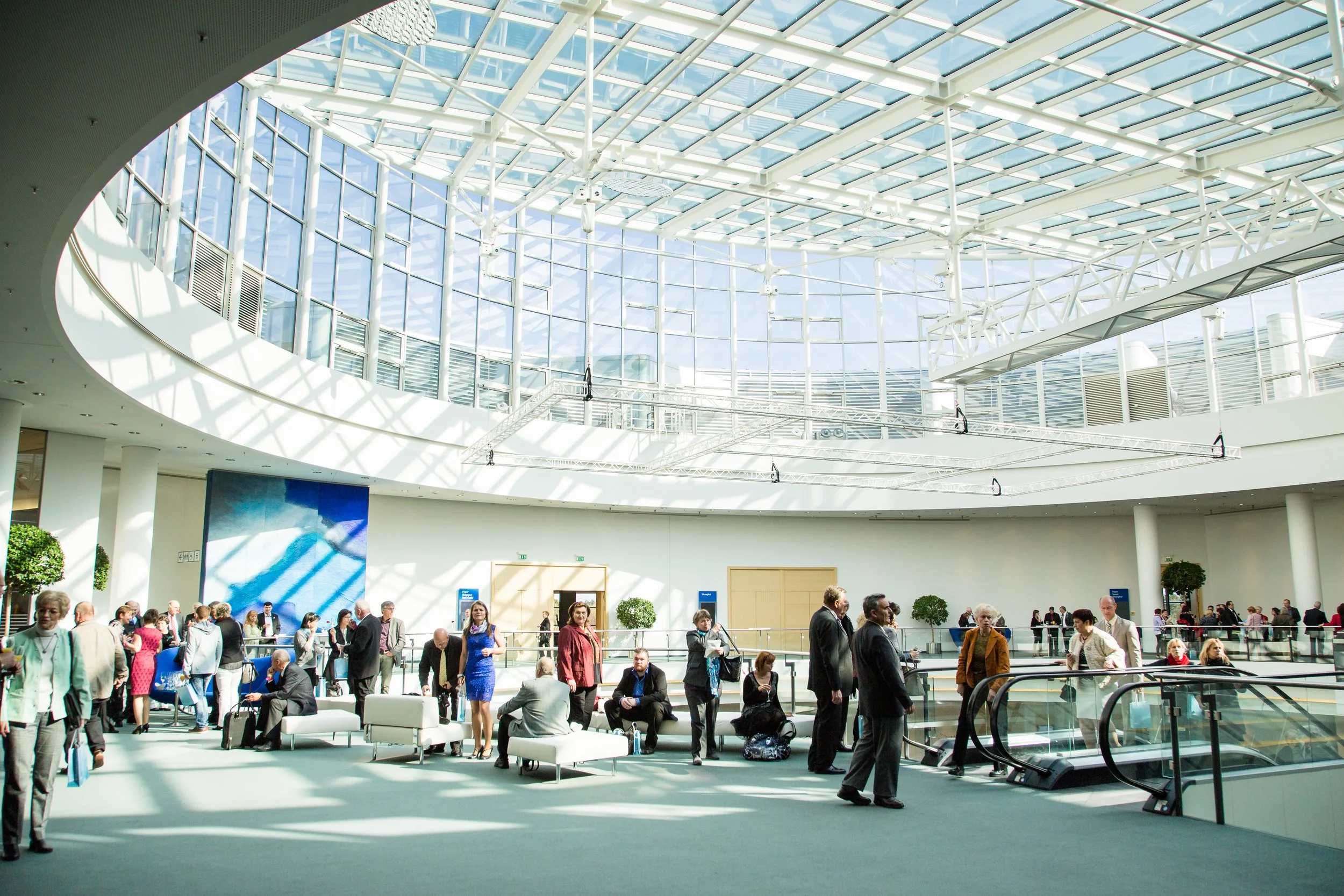 Innenansicht eines modernen Flughafens mit Glasdach, Warteschlangen, Sitzgelegenheiten und Menschen, die auf ihre Flüge warten.