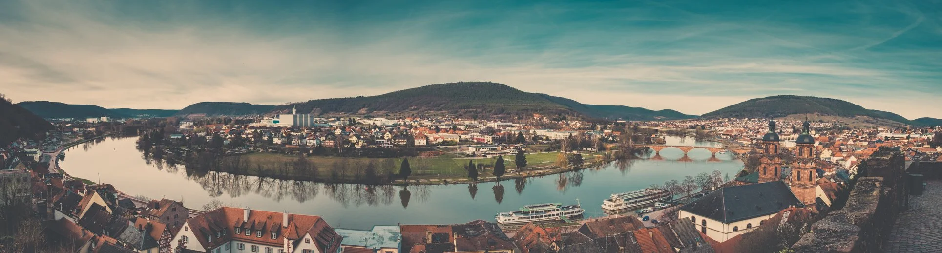 Panoramablick auf eine Stadt an einem Fluss mit Brücken, Kirchen und umgebenden Hügeln bei bewölktem Himmel.