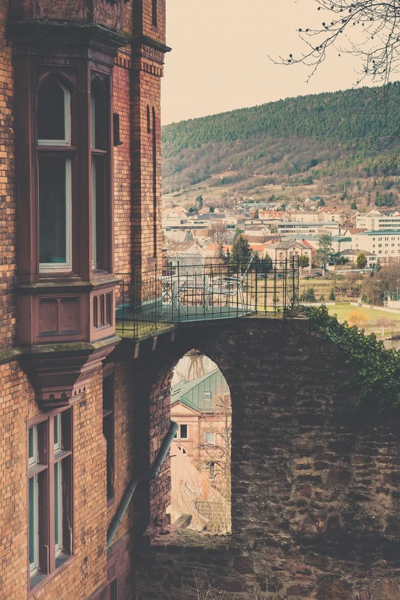 Ansicht eines historischen roten Backsteingebäudes mit hohen Fenstern und einer kleinen Terrasse am Rand, mit Blick auf eine Stadt und grüne Hügel im Hintergrund.