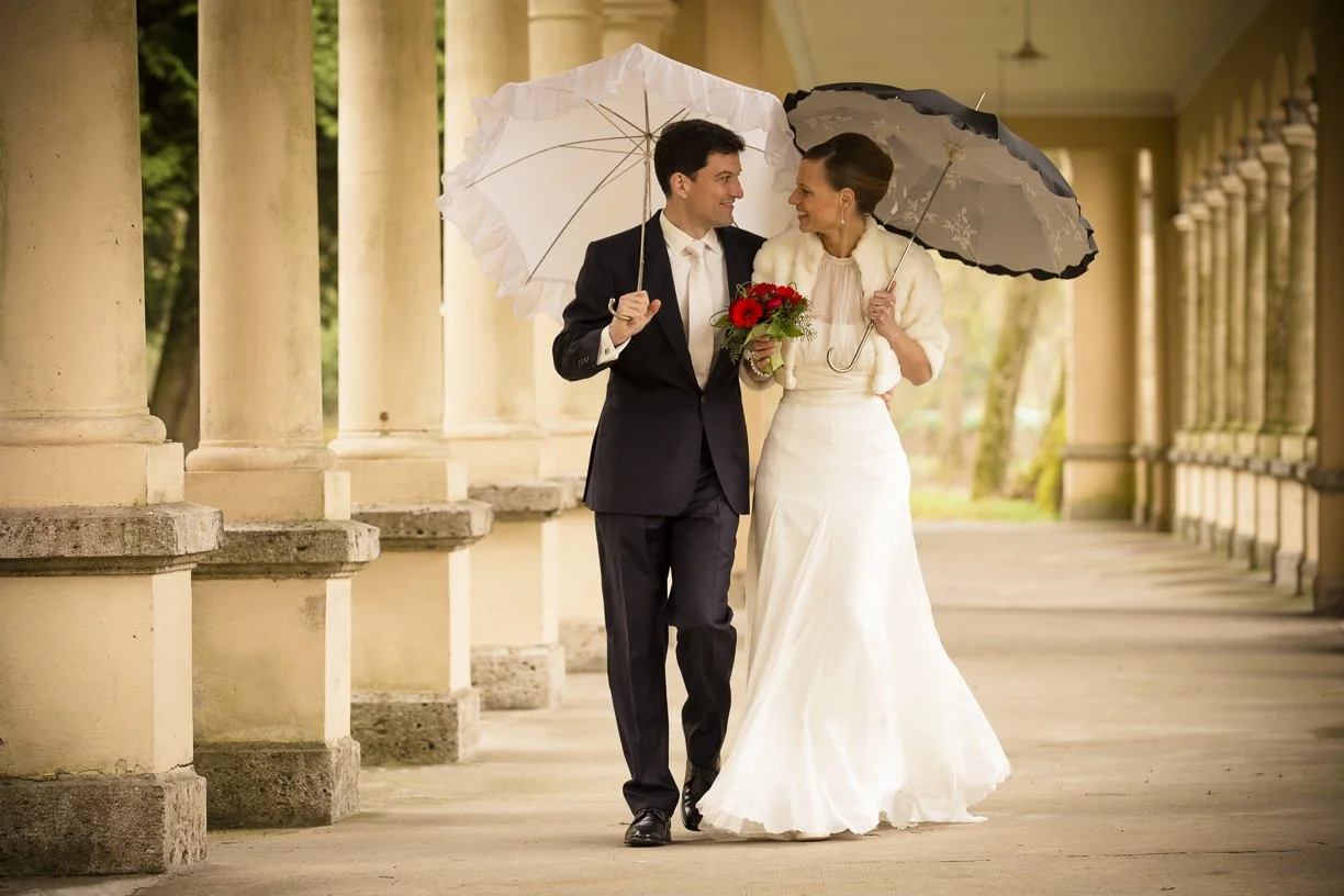 Ein Brautpaar im Hochzeitsoutfit geht arm in Arm durch eine Säulenhalle und hält Regenschirme, die sie vor Regen schützen, während sie sich liebevoll anschauen.