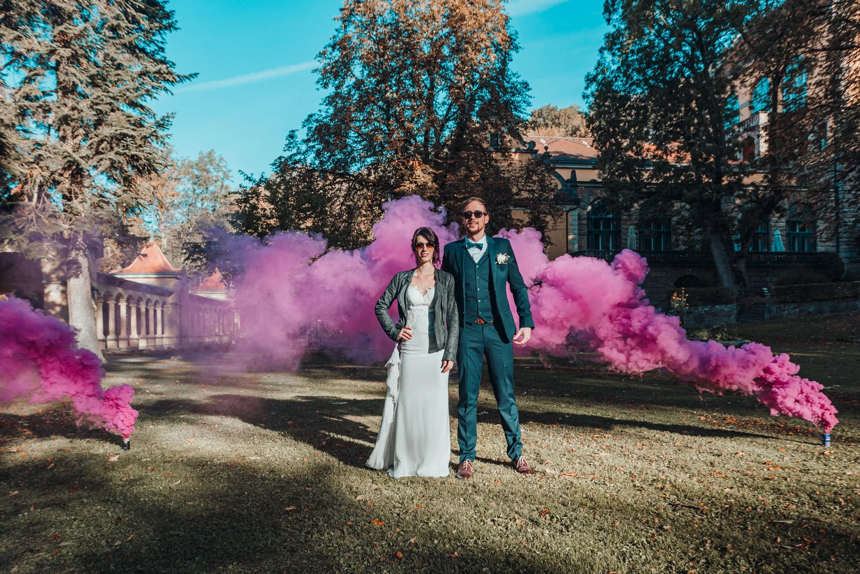 Ein Brautpaar steht im Freien bei sonnigem Wetter, im Hintergrund pinke Rauchschwaden, umgeben von Bäumen und einer historischen Villa.