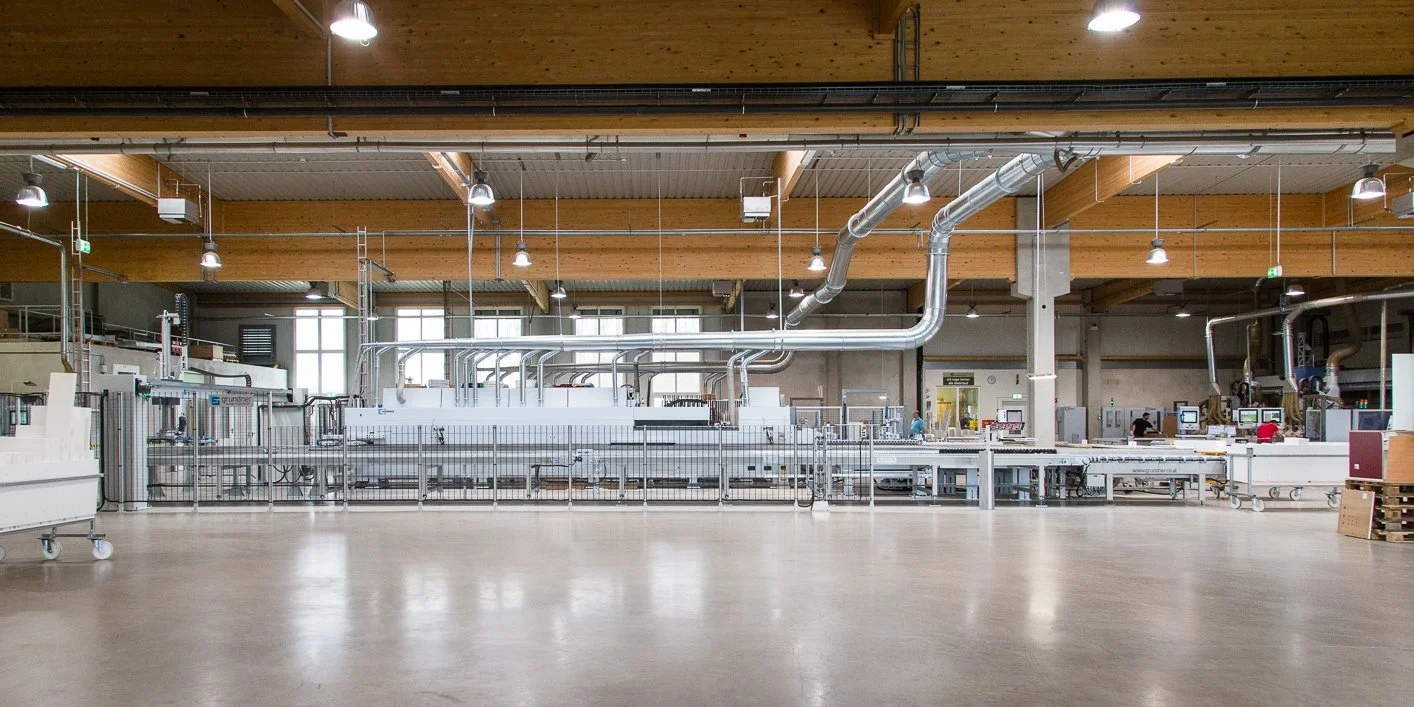 Große Industriehalle mit Produktionsmaschinen und technischen Anlagen, hellem Holzdecke, großen Fenstern und Beleuchtung, mit wenigen Mitarbeitern.