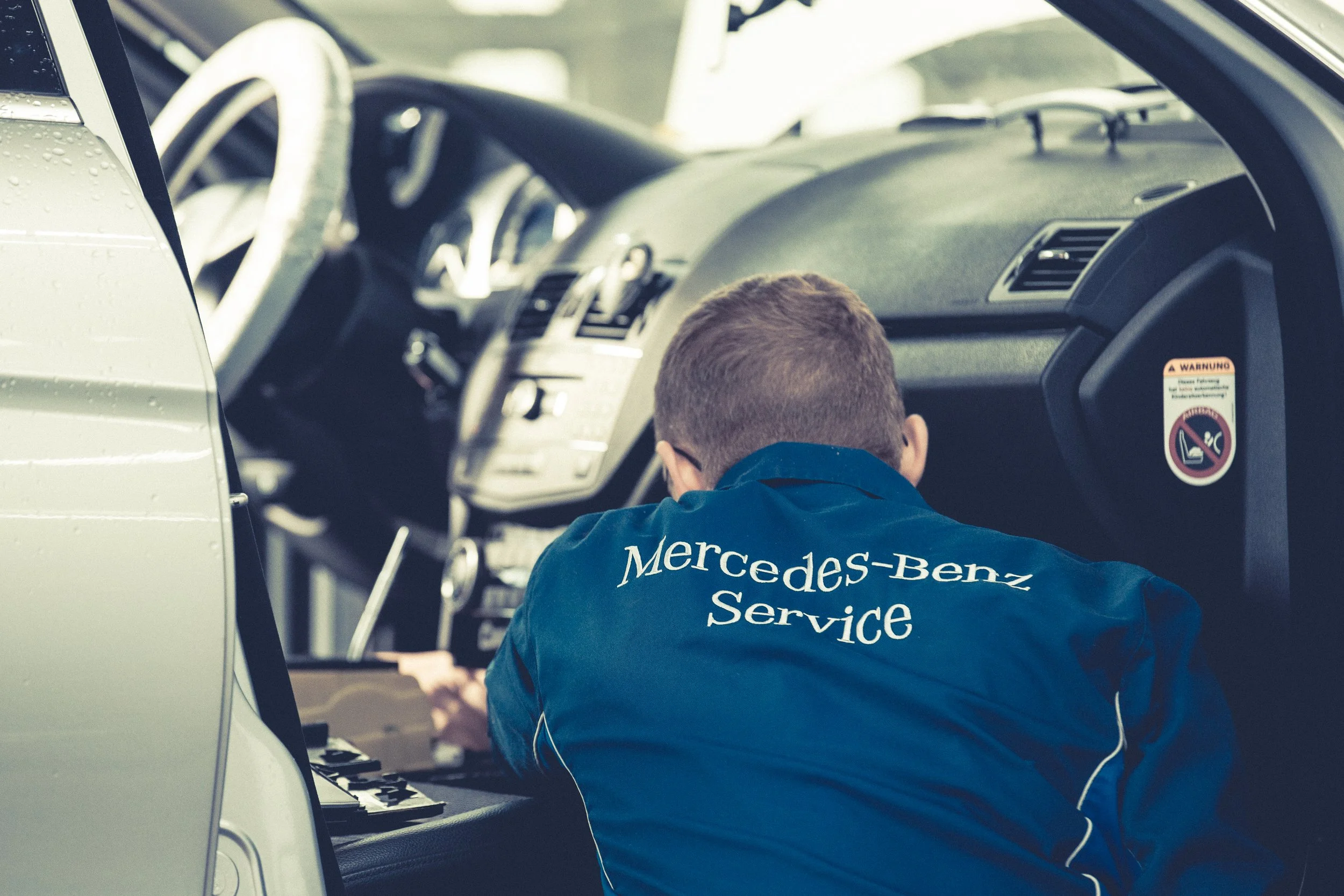 Ein Mann mit Mercedes-Benz Service Kleidung arbeitet im Inneren eines Autos, wahrscheinlich an Reparatur oder Wartung.