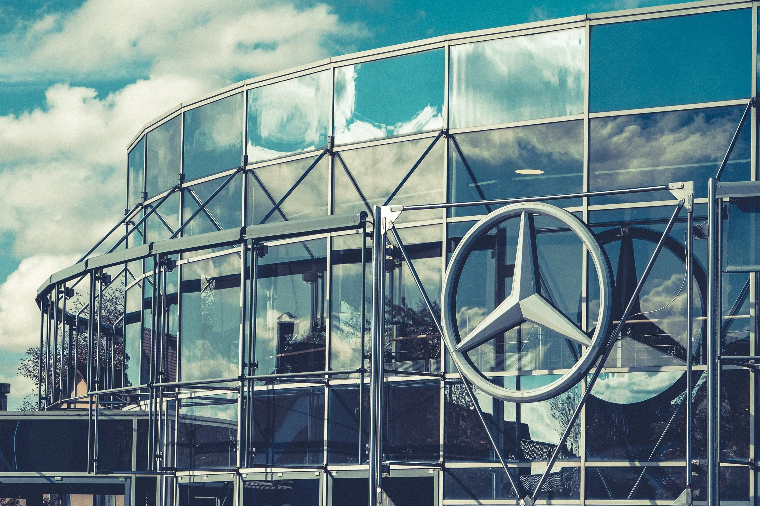 Ein modernes Gebäude mit großen Glasflächen und einem Mercedes-Benz Logo, reflektierende Fenster, blauer Himmel mit Wolken.
