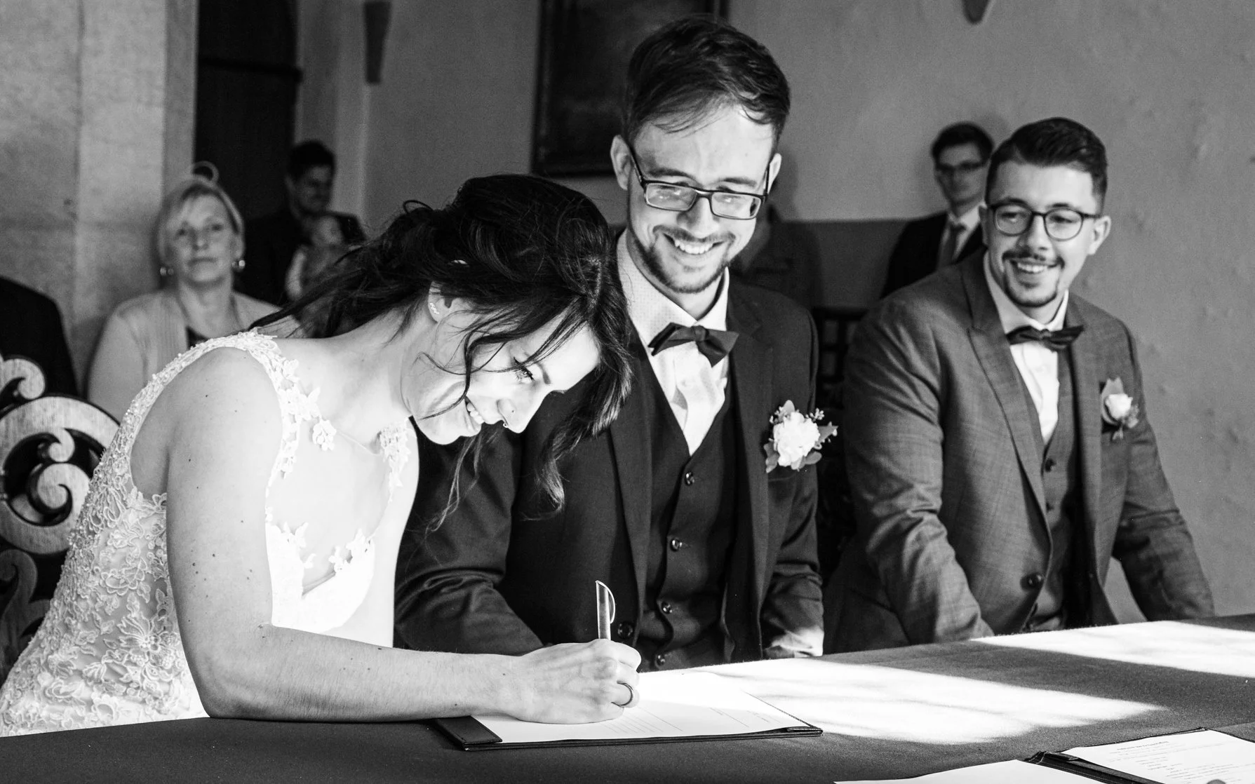 Braut und Bräutigam unterschreiben eine Urkunde bei einer Hochzeit während der Trauung, umgeben von Gästen in einem Raum mit gedämpfter Beleuchtung.