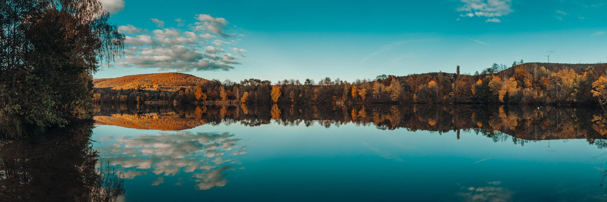 Eine ruhige Seenlandschaft im Herbst mit bunten Bäumen am Ufer, deren Spiegelbild im Wasser deutlich sichtbar ist, und einem blauen Himmel mit Wolken.