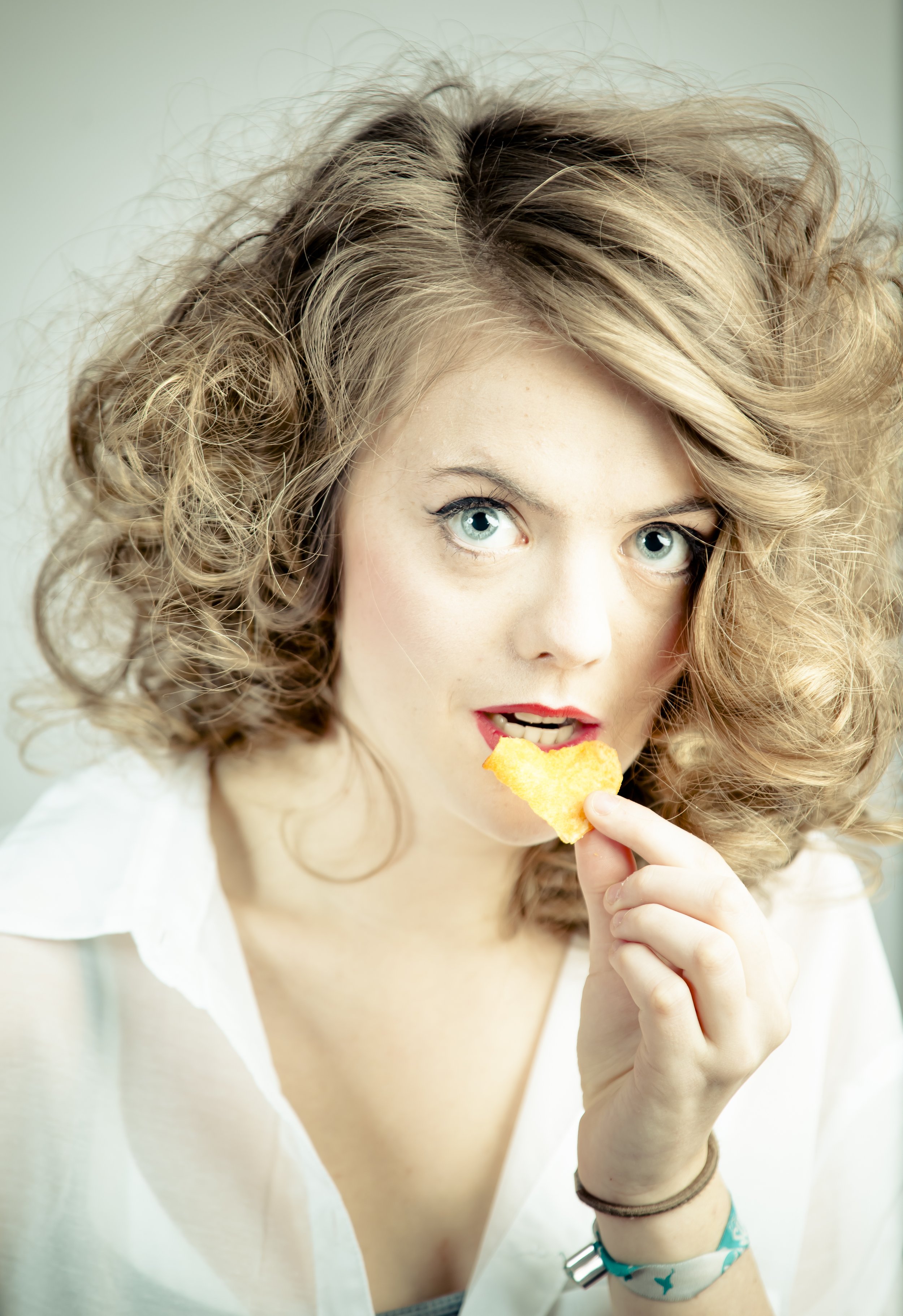 Junge Frau mit lockigem, blondem Haar, die einen Kartoffelchips hält und isst, trägt eine weiße Bluse und Armbänder.