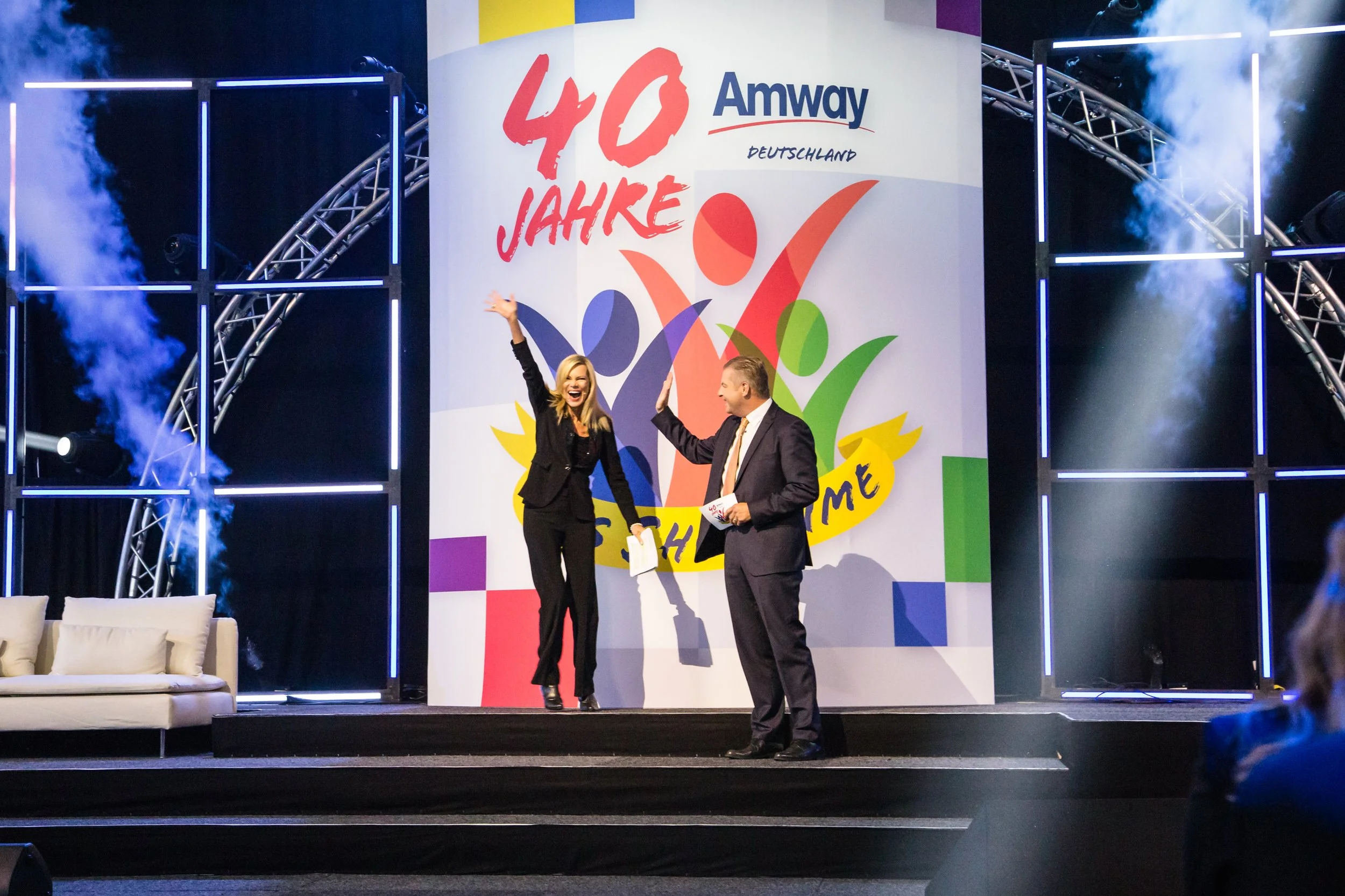 Zwei Menschen auf einer Bühne, eine Frau und ein Mann, feiern und geben sich die Hand, im Hintergrund ein großes Banner mit der Aufschrift '40 Jahre' und 'Amway Deutschland', dekorative Lichter und Rauch sind sichtbar.