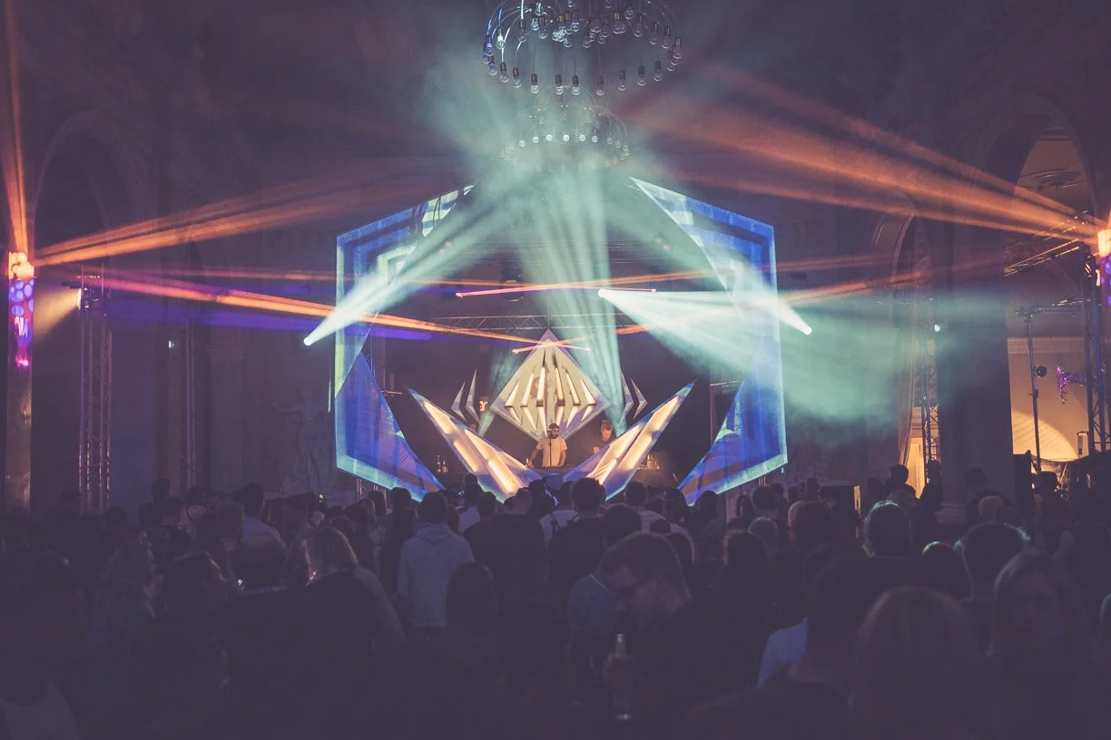 Musikveranstaltung mit DJ auf der Bühne, bunte Lichter und Publikum im dunklen Raum