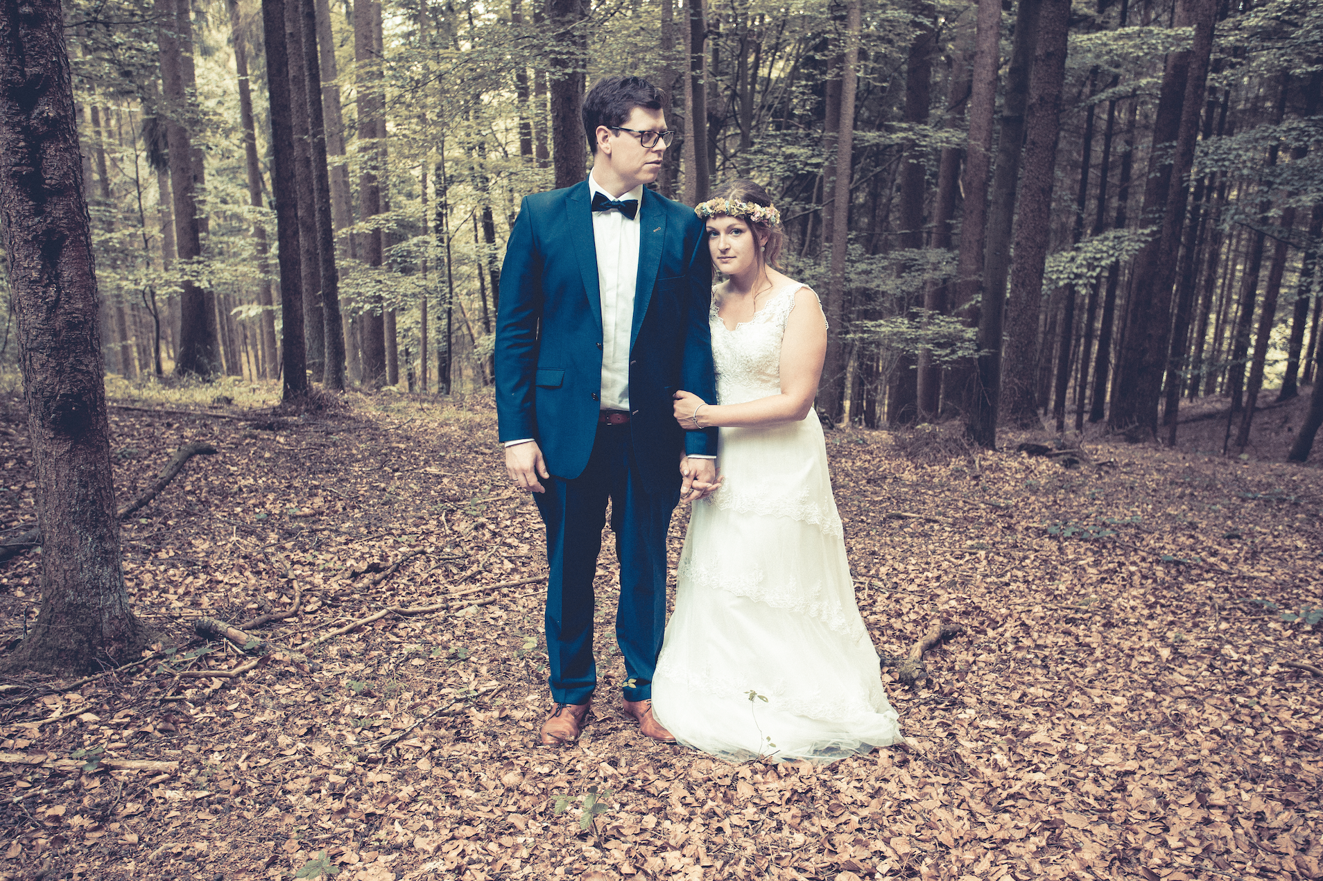 Ein Brautpaar steht in einem Wald. Der Bräutigam trägt einen blauen Anzug mit Fliege, die Braut ein weißes Hochzeitskleid mit Blumenkranz im Haar. Sie halten Händchen. Im Hintergrund sind viele Bäume.