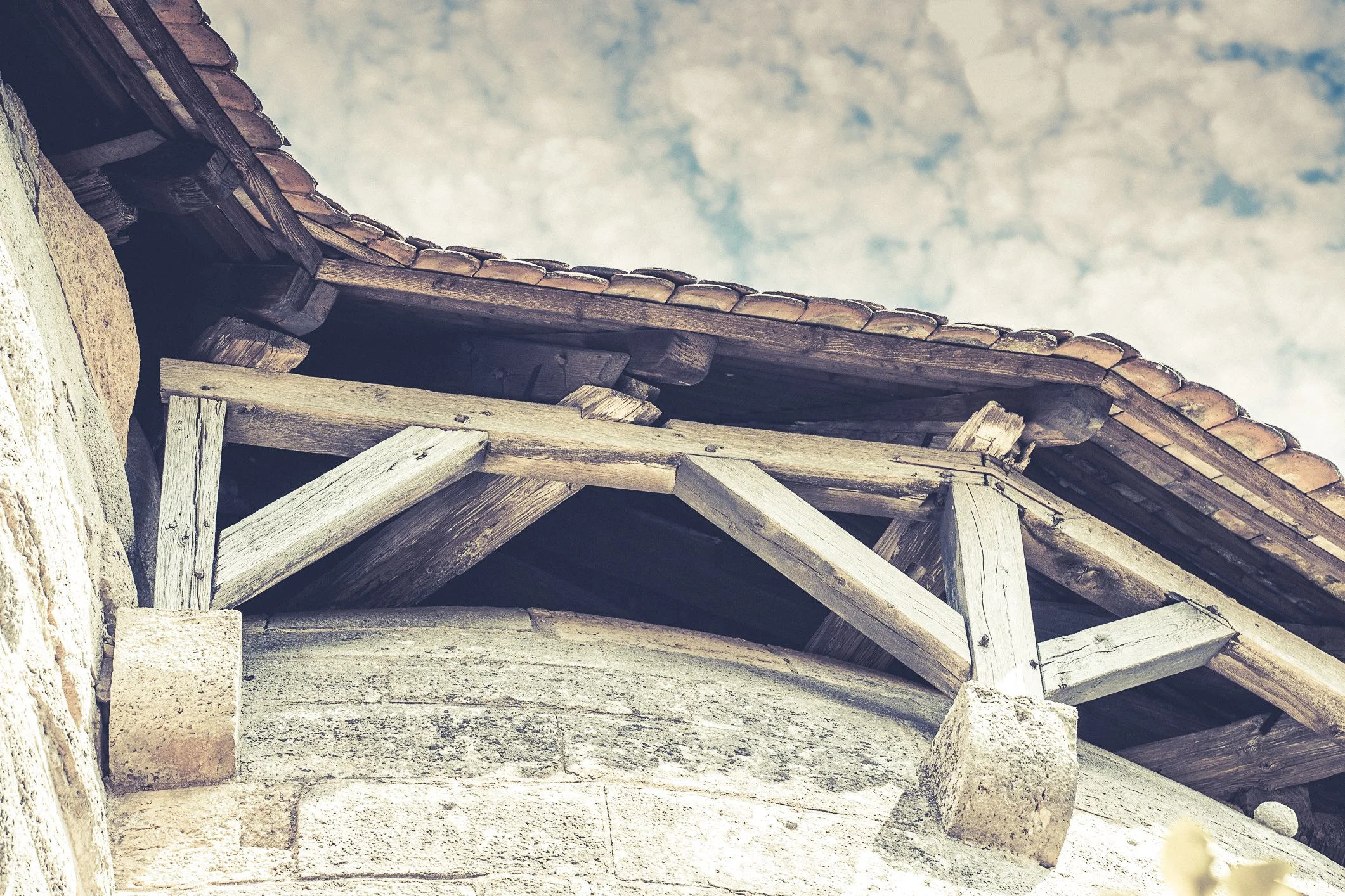 Ein alter Holzausbau an einer Steinwand mit einer Ziegeldachpartie darüber, Himmel im Hintergrund.