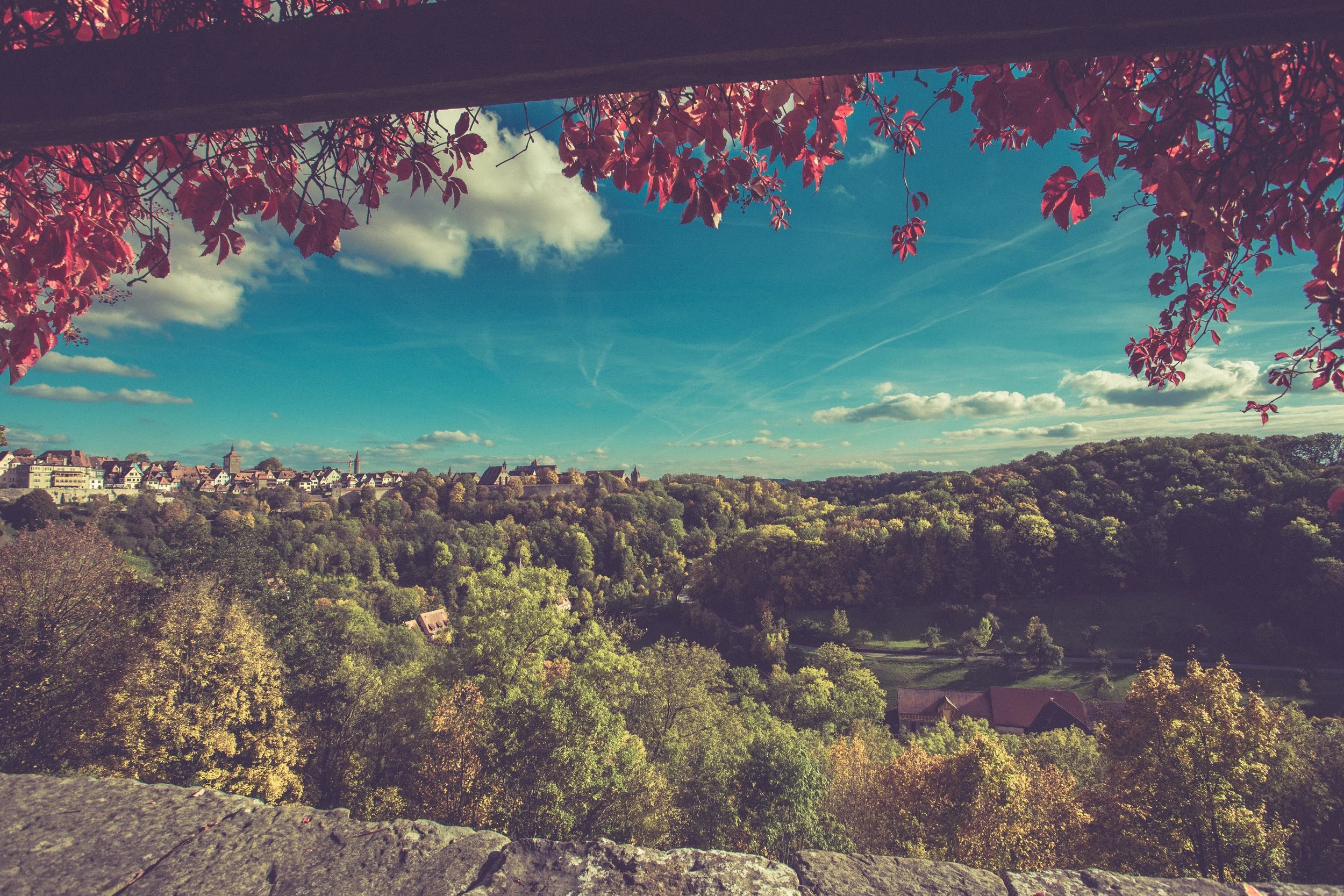 Blick auf eine ländliche Landschaft mit bunten Herbstbäumen, einem kleinen Dorf und einer Kirche im Hintergrund unter einem blauen Himmel mit Wolken.