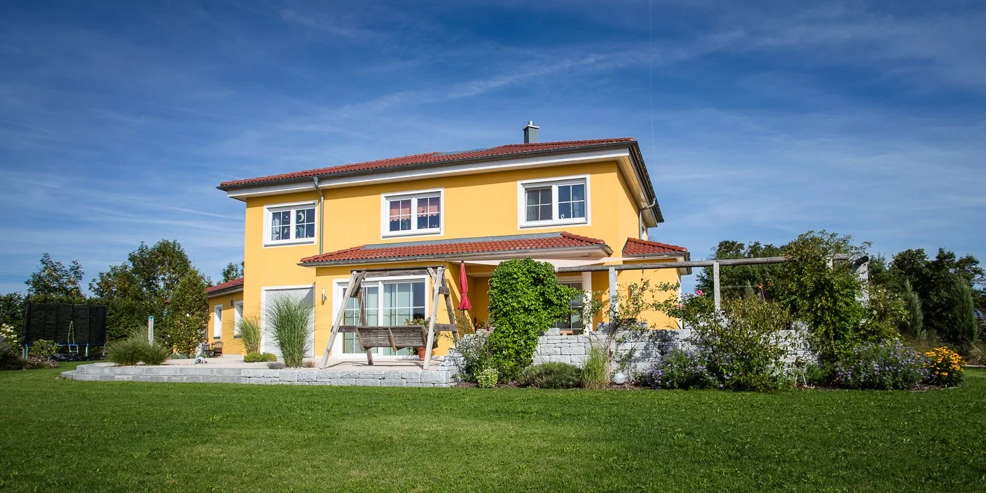 Ein zweistöckiges gelbes Haus mit roten Ziegeldach, umgeben von einem grünen Garten mit Rasen, Pflanzen und Bäumen.