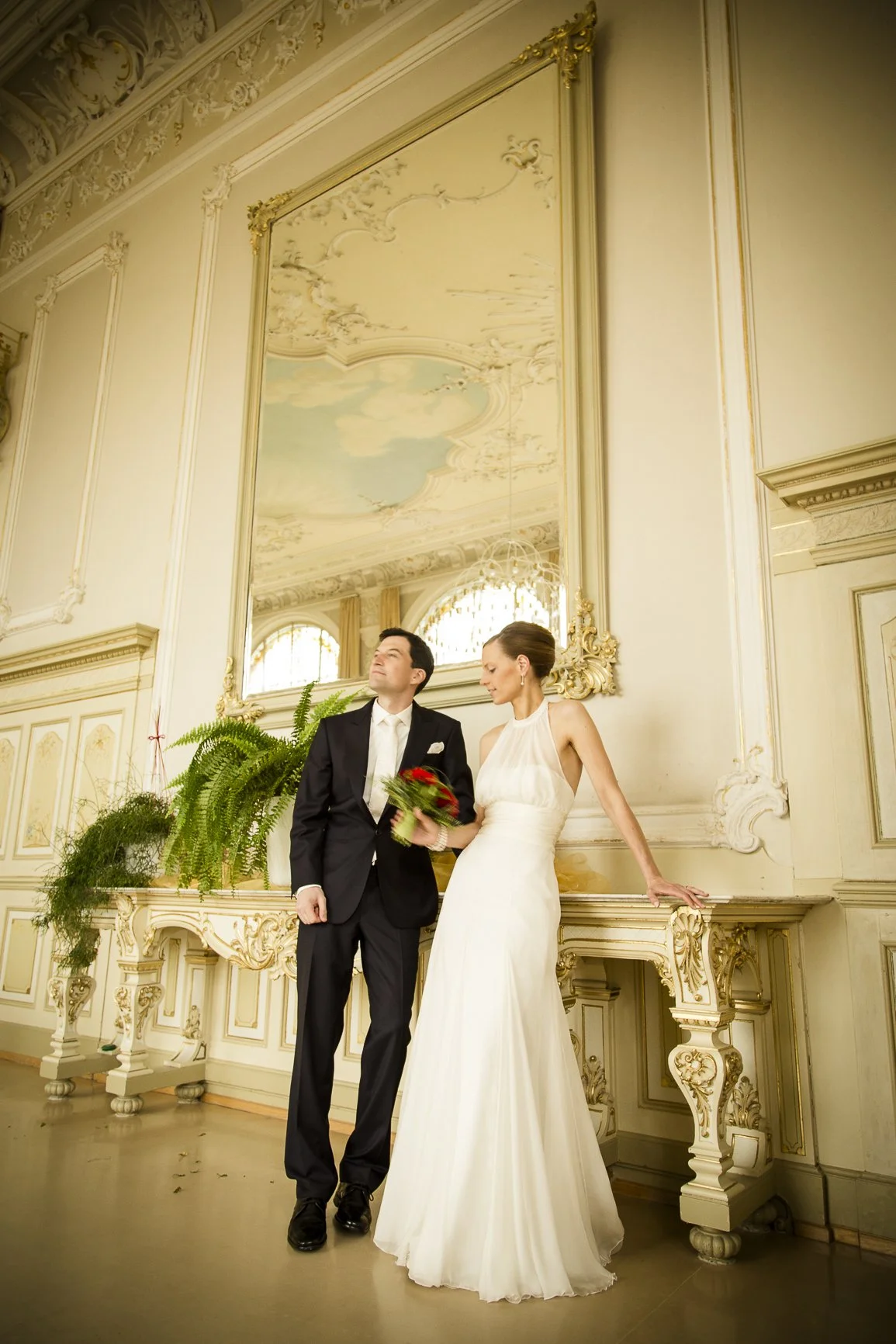 Ein frisch verheiratetes Paar steht in einem eleganten, barocken Raum. Der Bräutigam trägt einen schwarzen Anzug mit weißem Hemd, die Braut trägt ein weißes Hochzeitskleid und hält einen Blumenstrauß in der Hand.