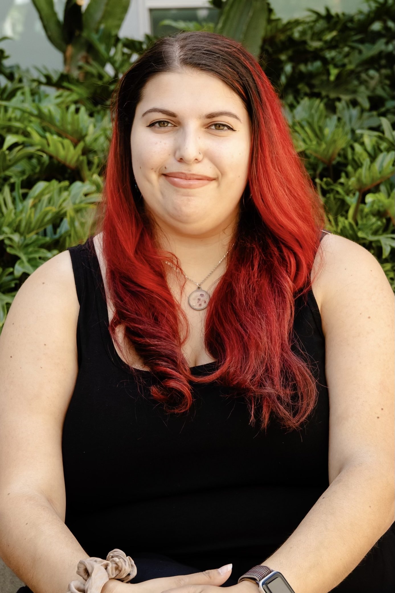 Headshot of Lexi, smiling with wavy red hair, wearing a black shirt, with green plant leaves in the background.