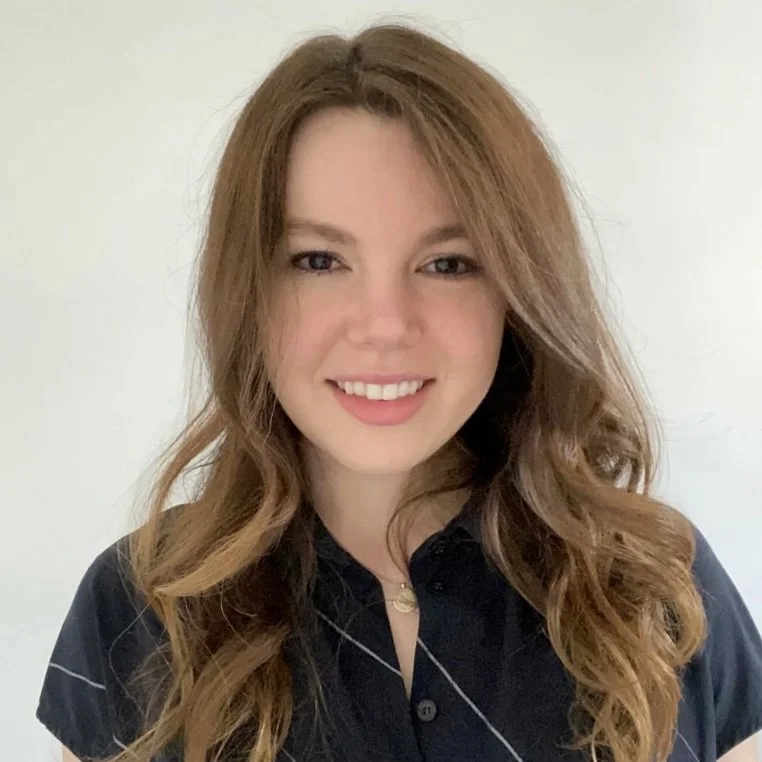 Headshot of Claire, smiling with wavy brunette hair, wearing a dark blue shirt against a white background.