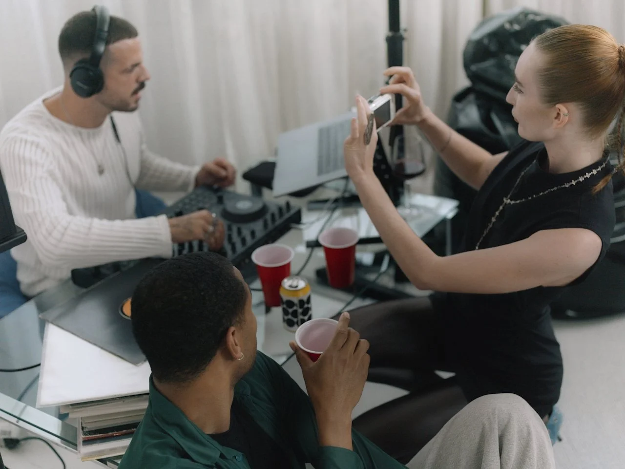 A DJ working at a mixing setup while two friends record and take photos behind the scenes at a casual studio gathering.