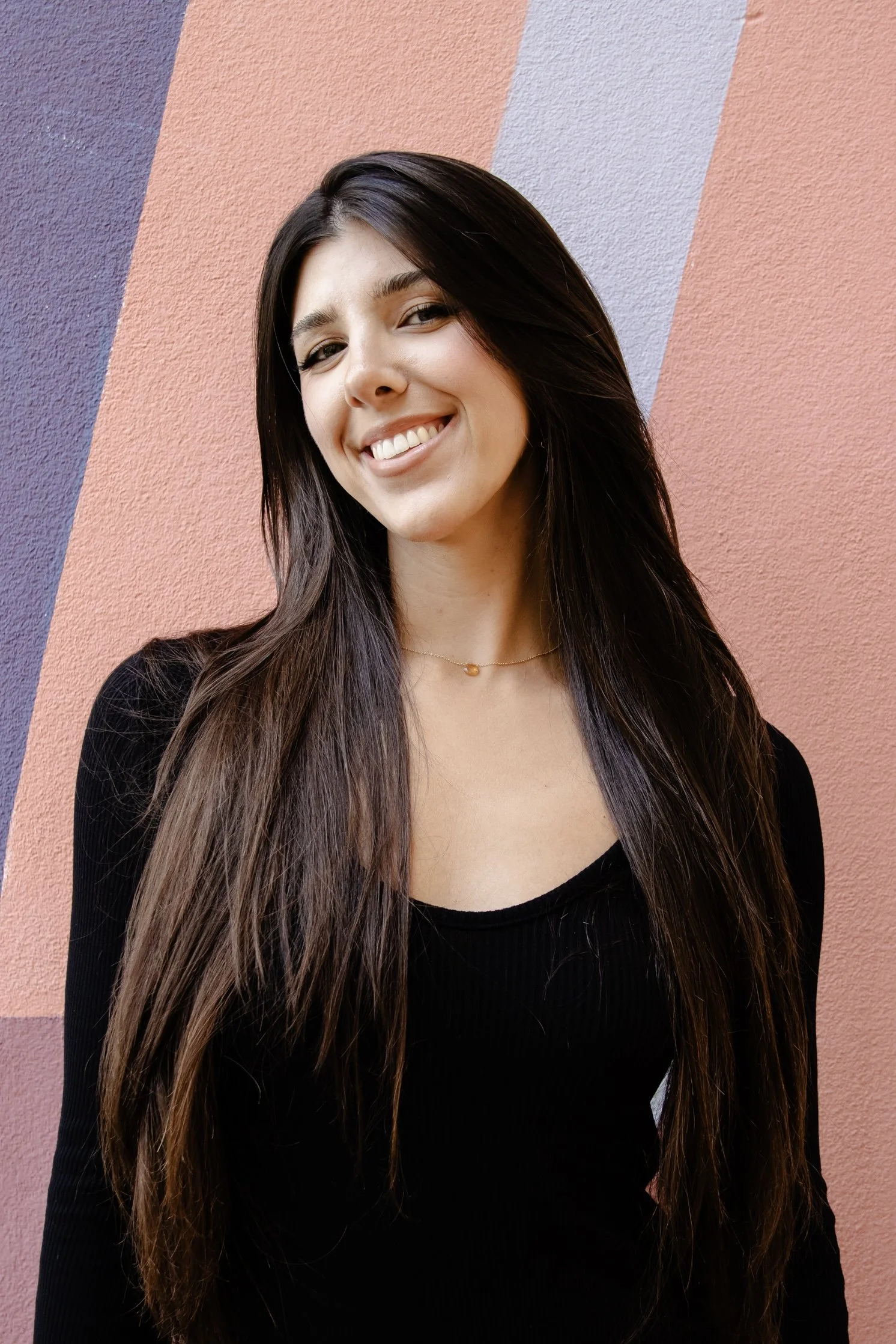 Headshot of Gabby standing in front of a colorful geometric background, smiling with long dark hair and wearing a black top.”