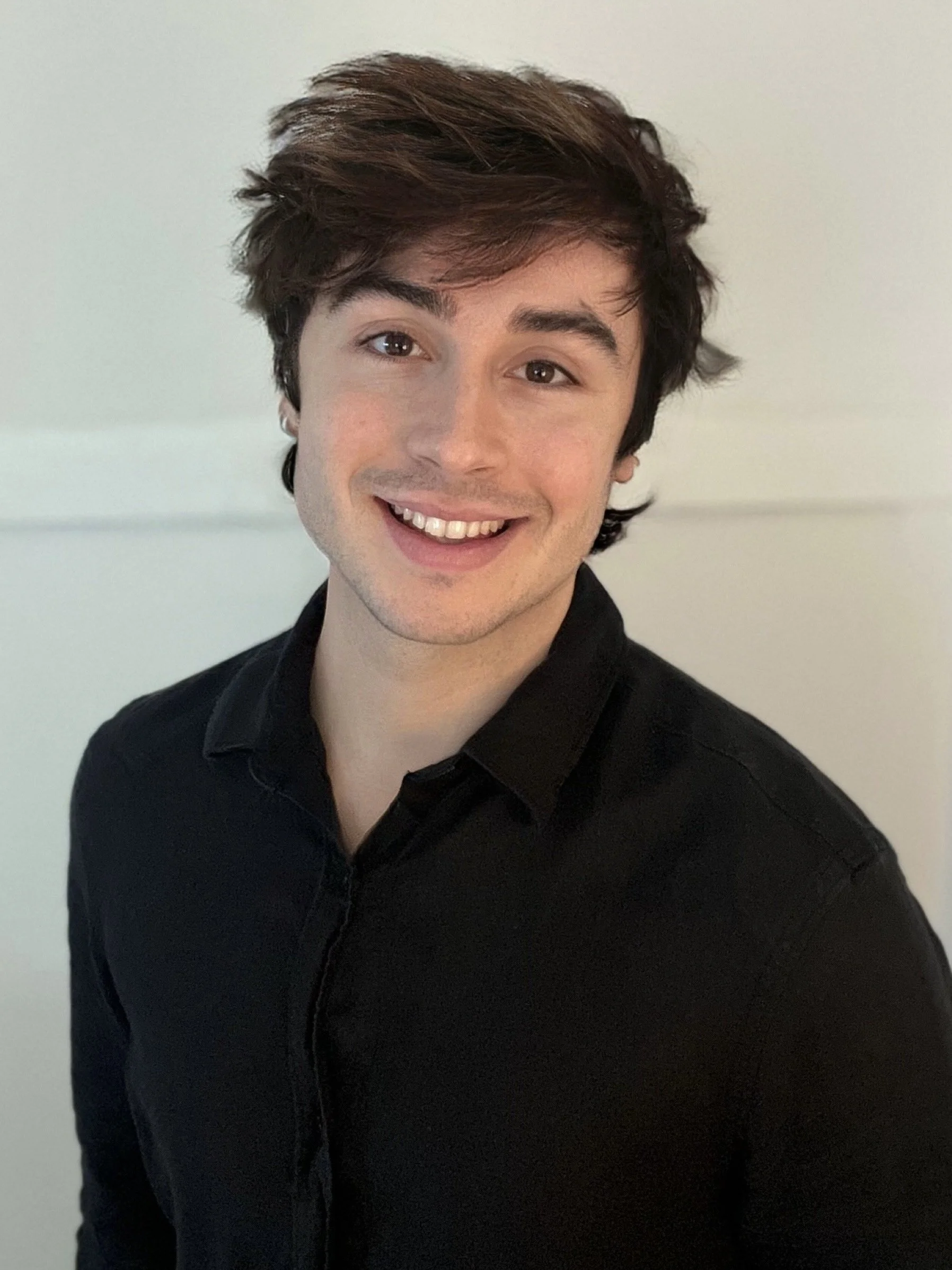 Headshot of Matthew, smiling with medium-length dark hair stands against a light background, wearing a black collared shirt.