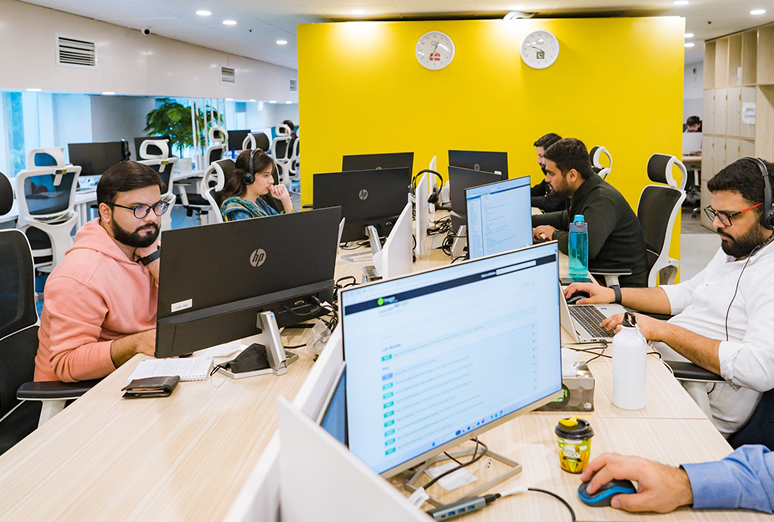 Employees working at computers in an open office with a bright yellow accent wall and clocks showing different time zones